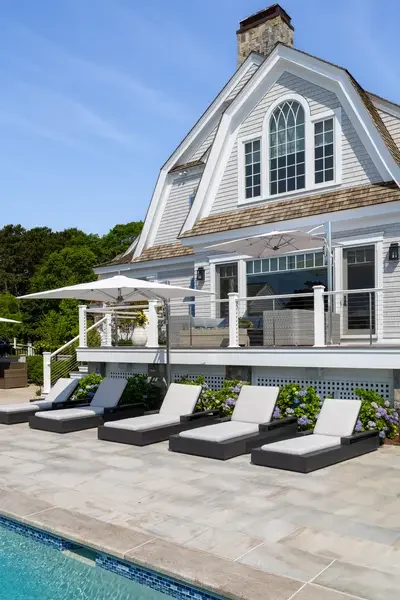 A house with a large deck area featuring multiple lounge chairs, an outdoor umbrella, and a swimming pool in the foreground.