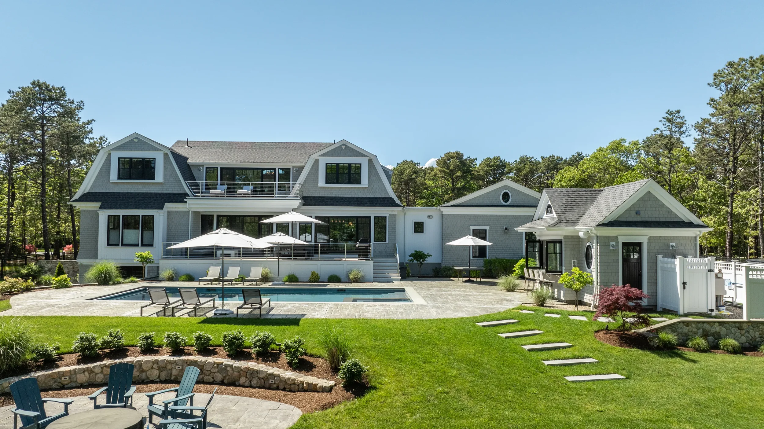 A luxurious backyard with a swimming pool, lounge chairs, umbrellas, a landscaped garden, and a large modern house with multiple stories and a deck, set against a backdrop of trees.