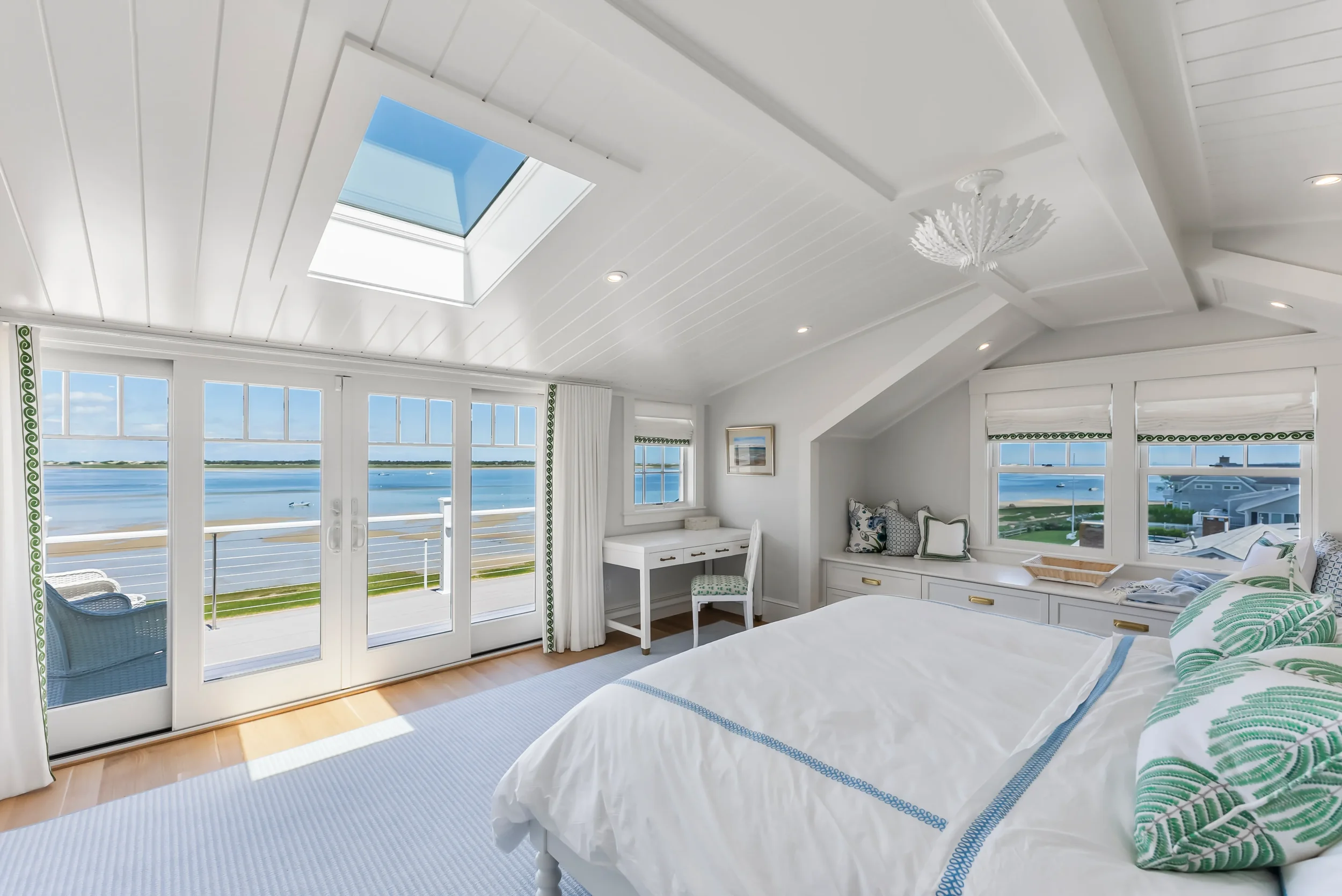Bright bedroom with large windows and a skylight, overlooking a body of water. The room has white walls and ceiling, decorated with pillows, a small desk, and a chair. A balcony with outdoor seating is visible through the sliding glass doors.