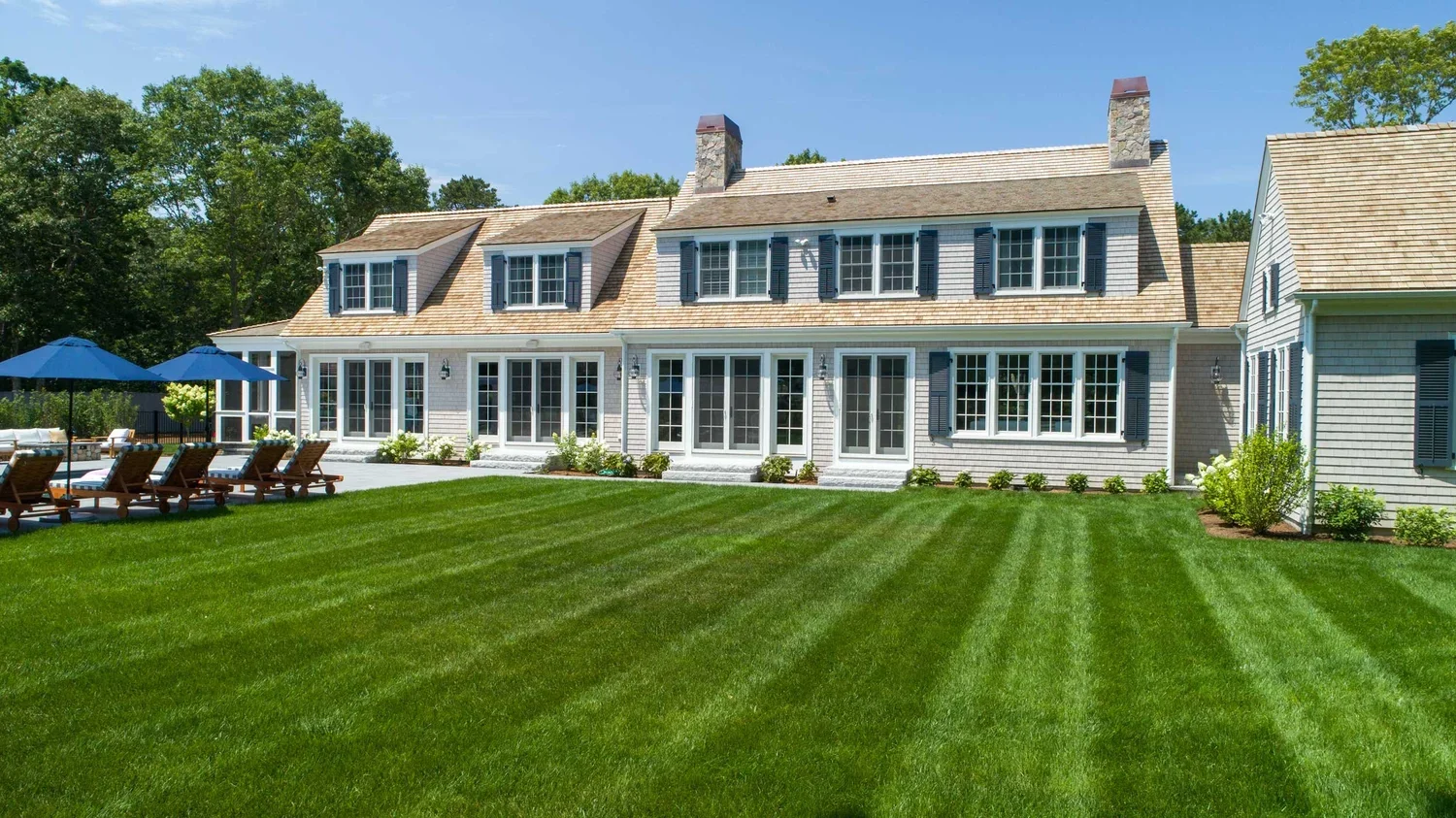 Large white house with multiple windows, black shutters, and a patio with lounge chairs and umbrellas in a grassy yard.