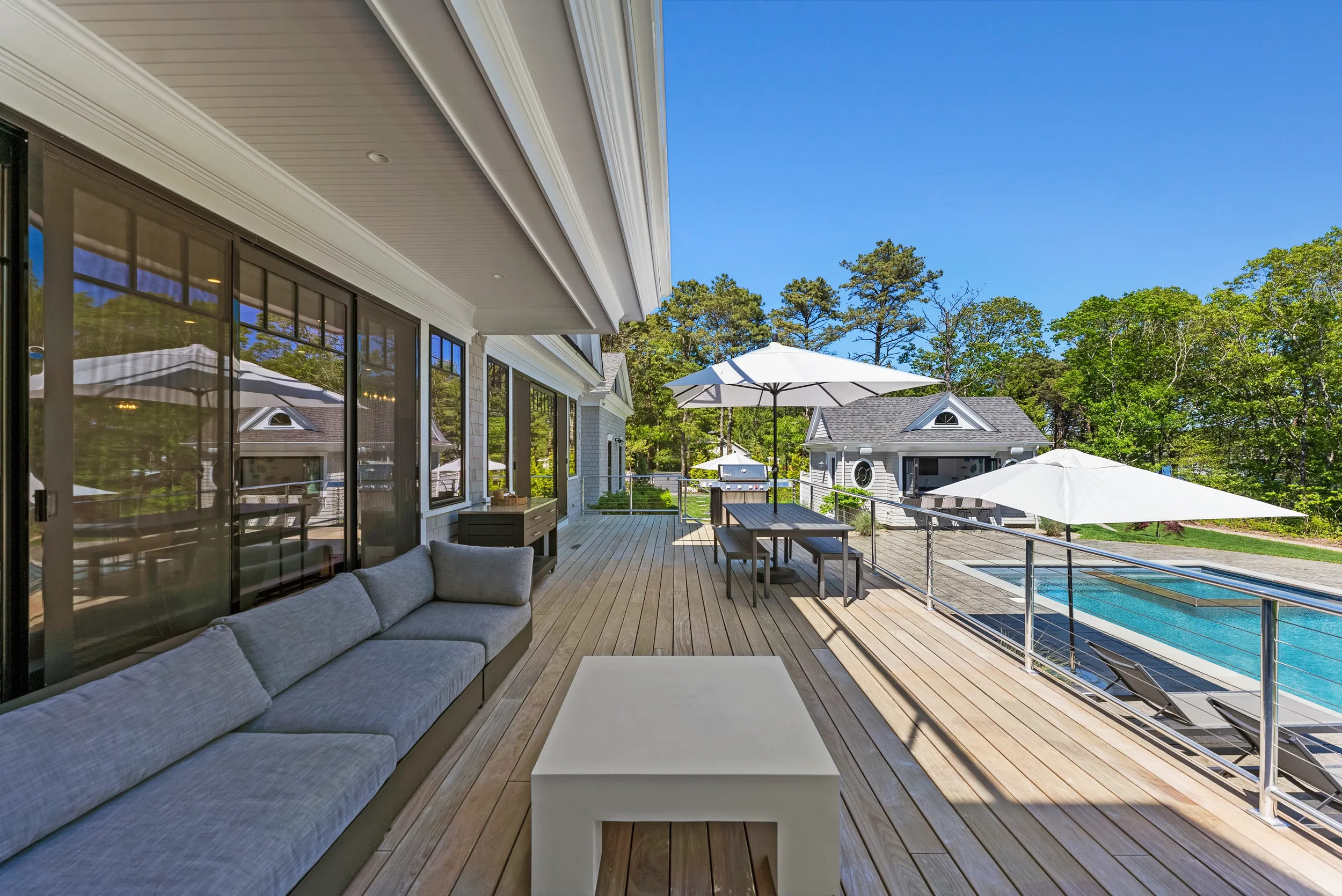 Large outdoor deck with gray sofa, white table, dining table with benches, umbrellas, and a swimming pool, surrounded by trees and neighboring houses.