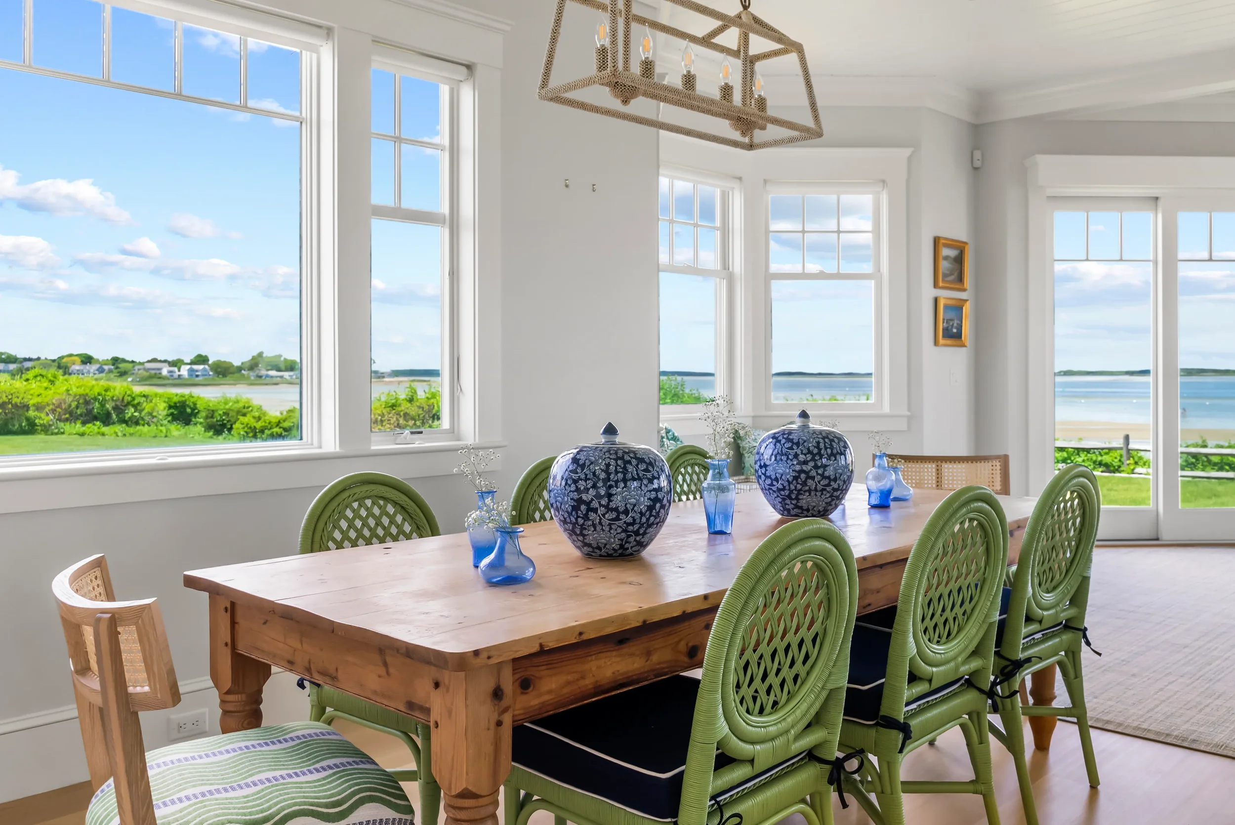 Bright dining room with large windows showcasing a scenic ocean view, a wooden table with blue and white vases, and green chairs.