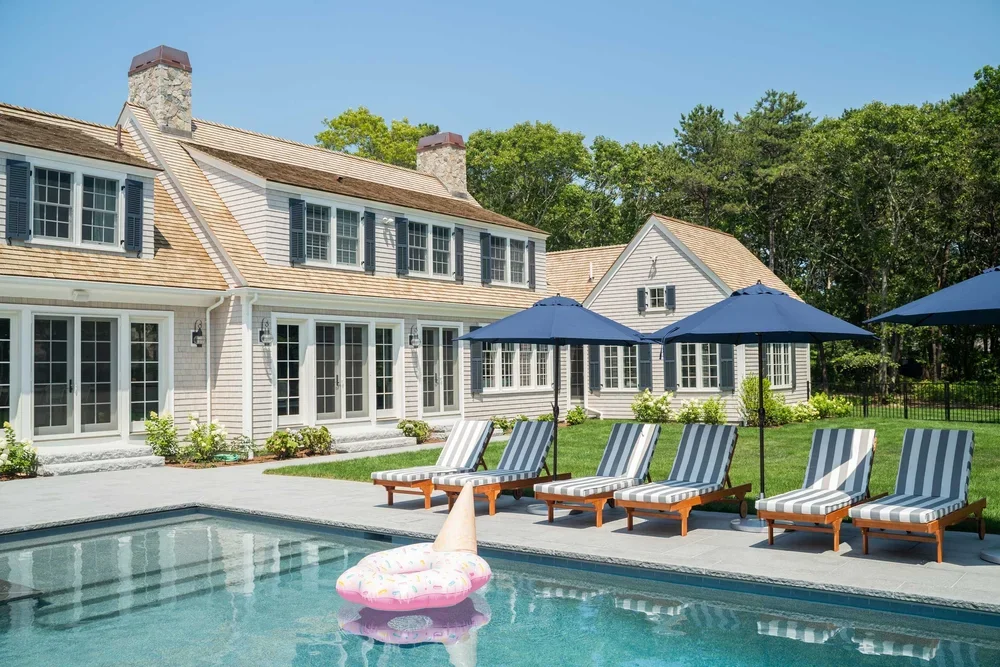 Backyard with a swimming pool, striped lounge chairs, blue umbrellas, and a large house with multiple windows and a chimney, surrounded by green trees.