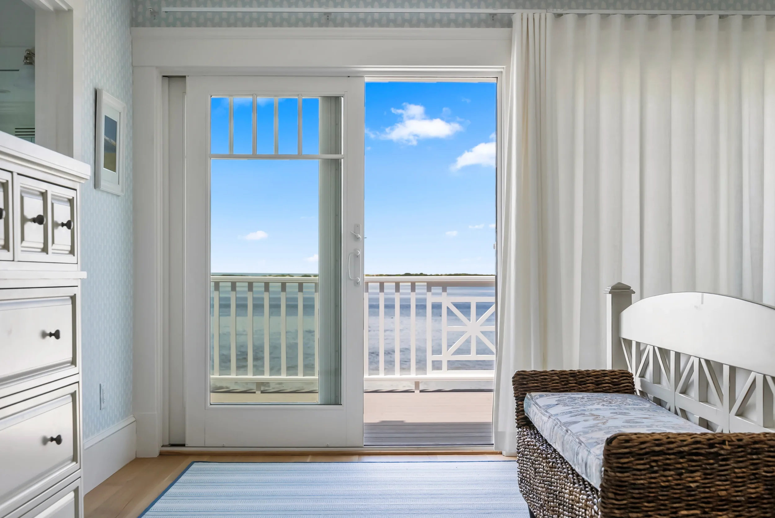 Room with sliding glass door leading to a balcony overlooking water and sky, wicker bench with cushion, white curtains, and light-colored furnishings.