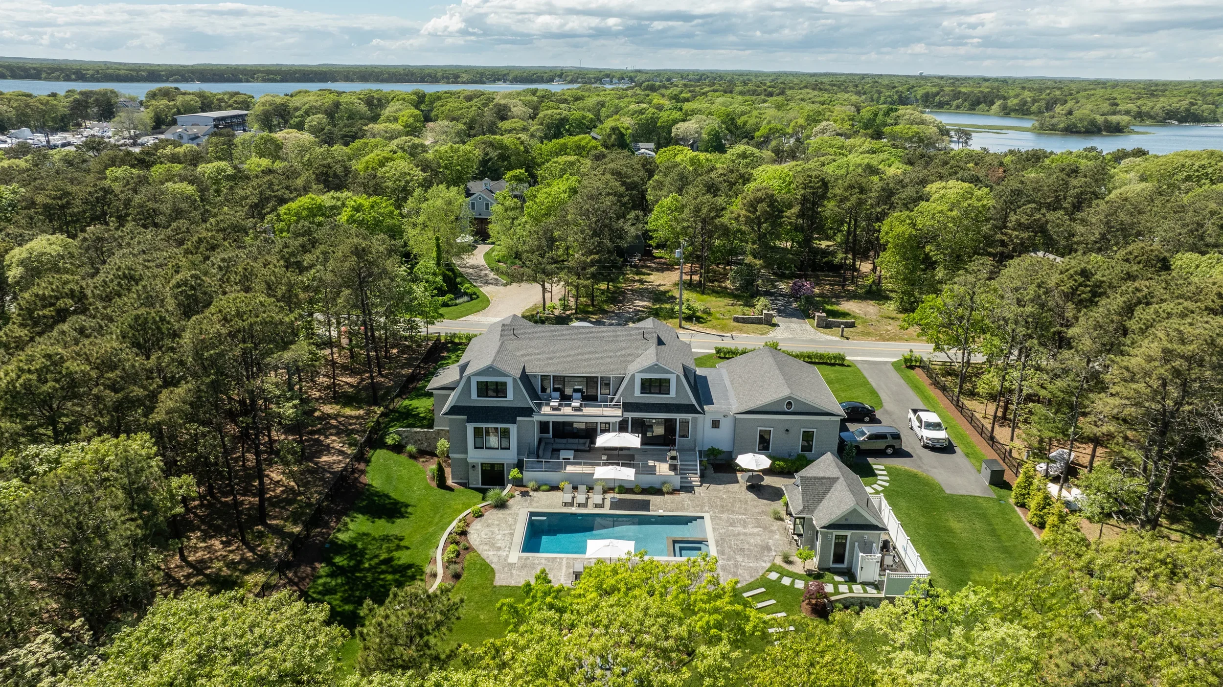 A large, modern house with a swimming pool and outdoor seating area, surrounded by trees and greenery, with a lake in the background.