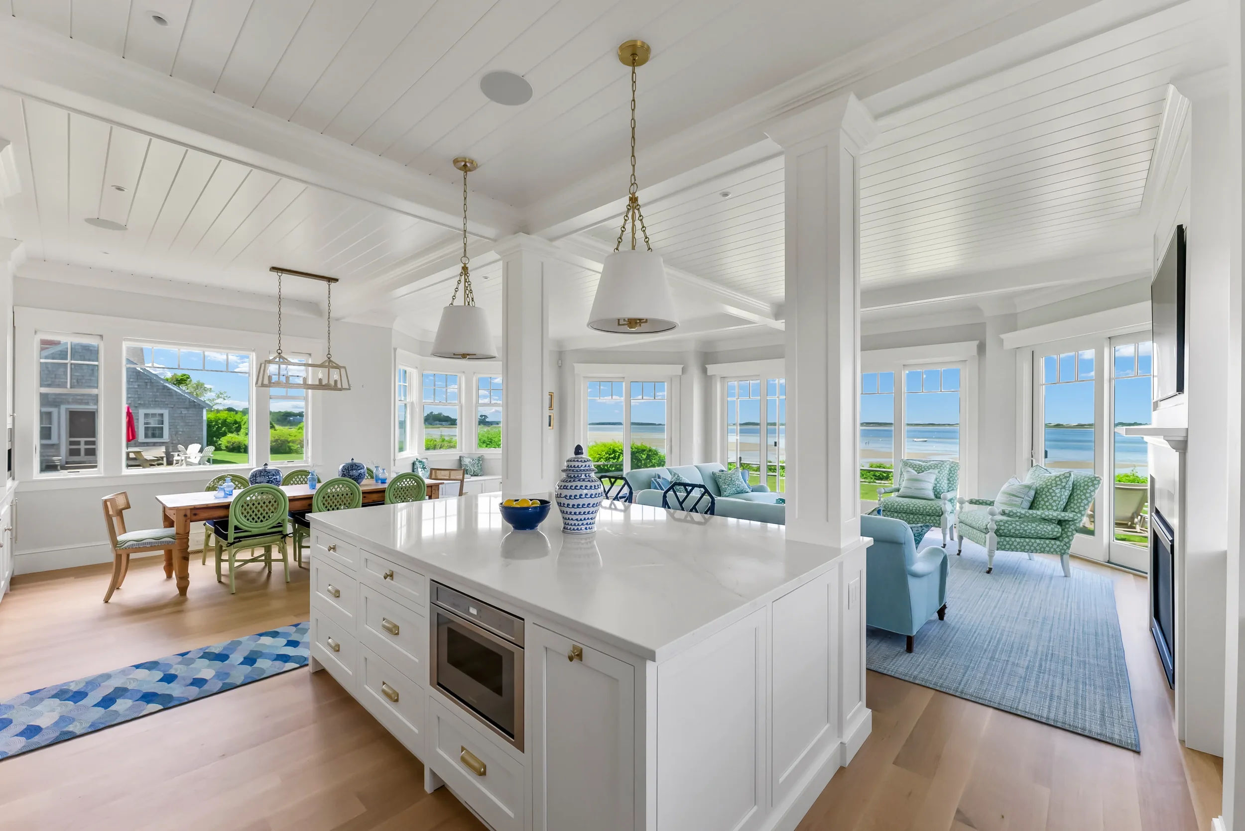 Bright, open living room and kitchen with large windows overlooking a lake, white walls, ceiling, and cabinetry, light wood floors, and coastal-themed furniture and decor.
