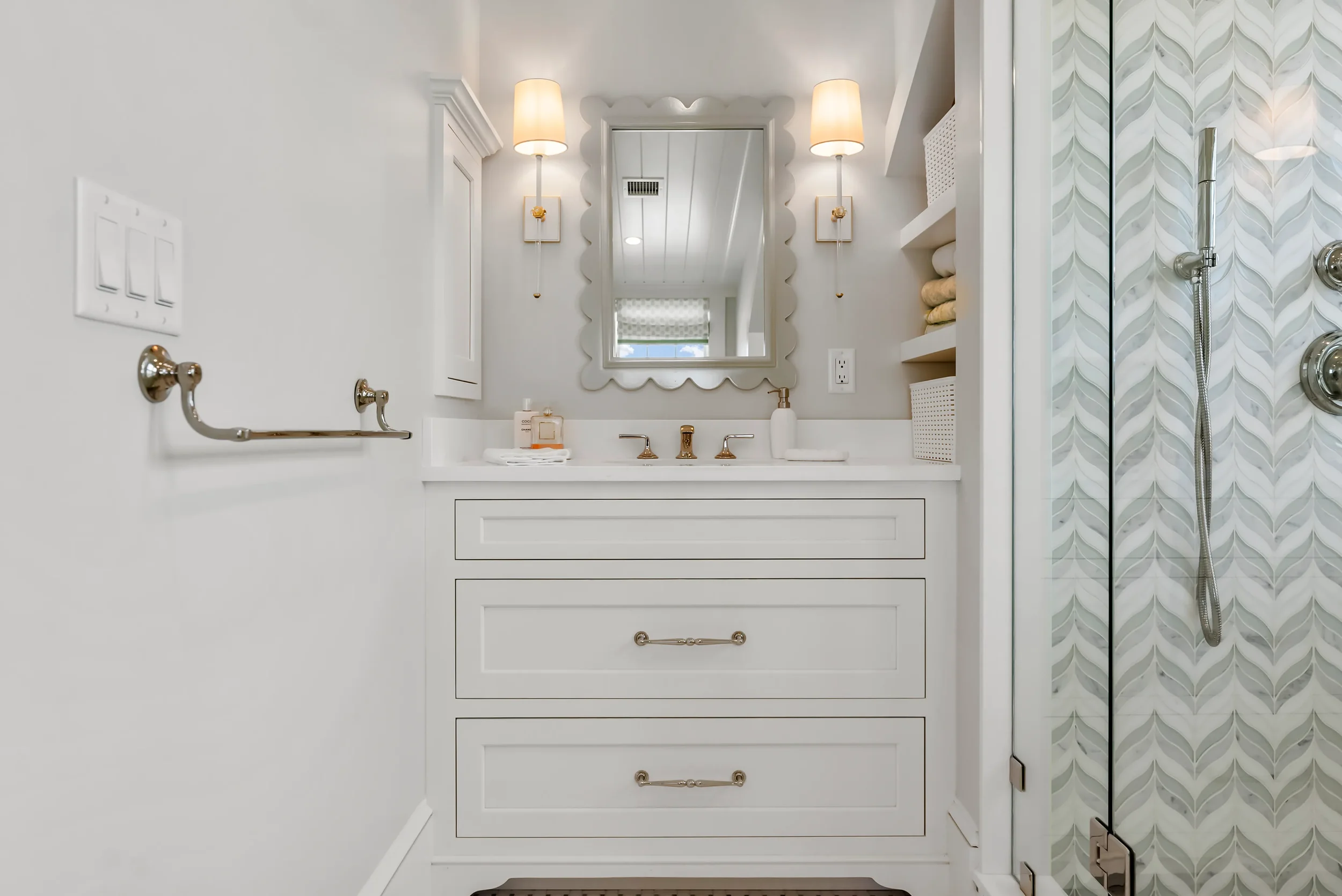 A white bathroom vanity with a mirror above it, two wall-mounted lamps flanking the mirror, and shelves with towels and baskets next to a shower with a glass door and patterned tile.