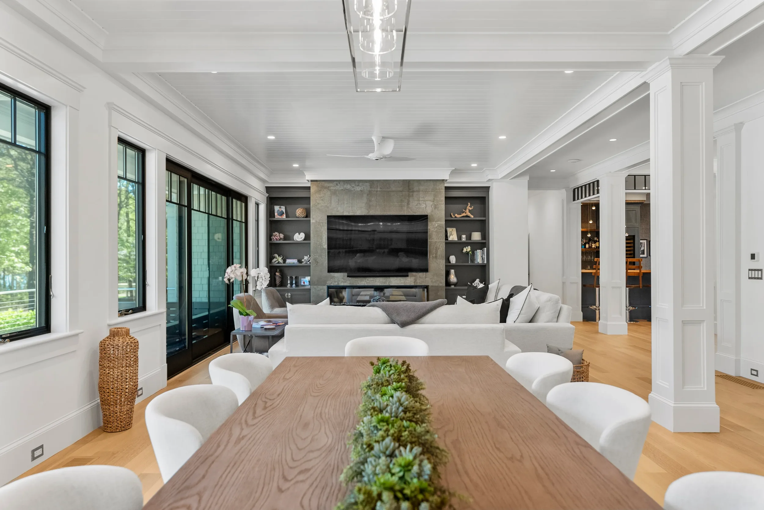 Modern open-concept living room and dining area with white walls, light wood floors, large windows, a wall-mounted TV, built-in shelves, and a wooden dining table with white chairs.
