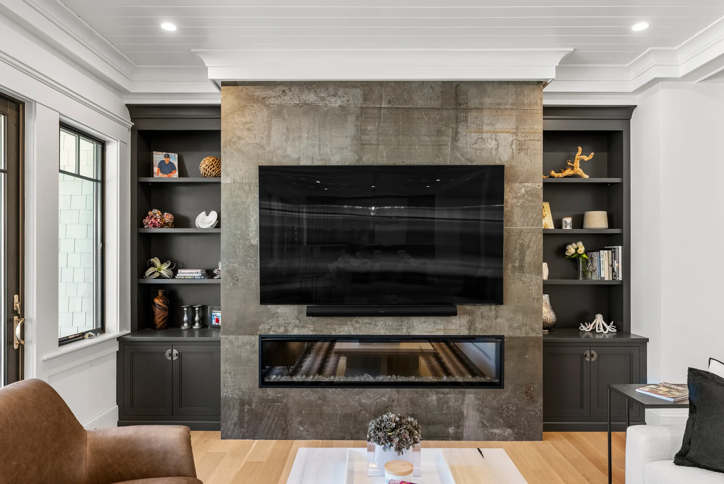 Living room with a large wall-mounted TV, modern fireplace, dark shelves on either side with decorative items and books, window on the left, and small tables with decor in the foreground.