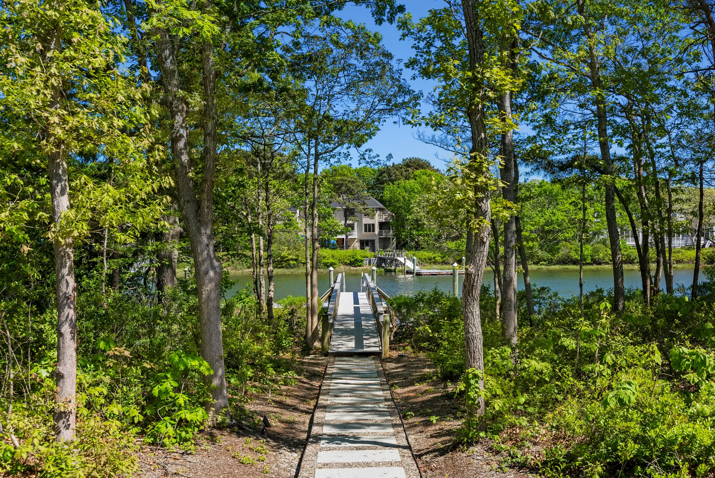 A narrow wooden walkway leads to a dock over a lake, surrounded by trees and greenery, with houses visible in the background under a clear blue sky.
