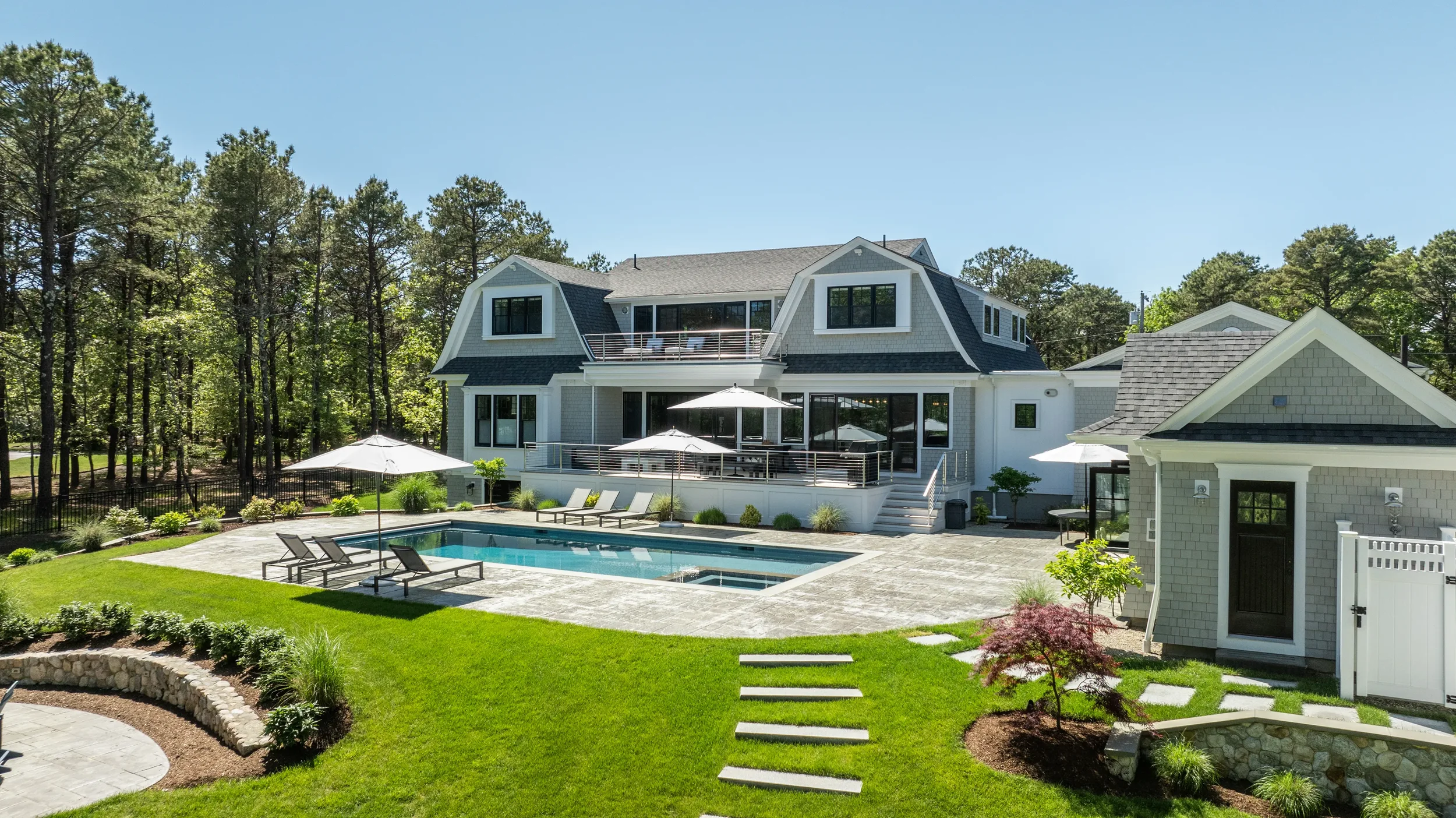 A modern house with a backyard featuring a swimming pool, lounge chairs, umbrellas, and landscaped lawn with trees and plants.