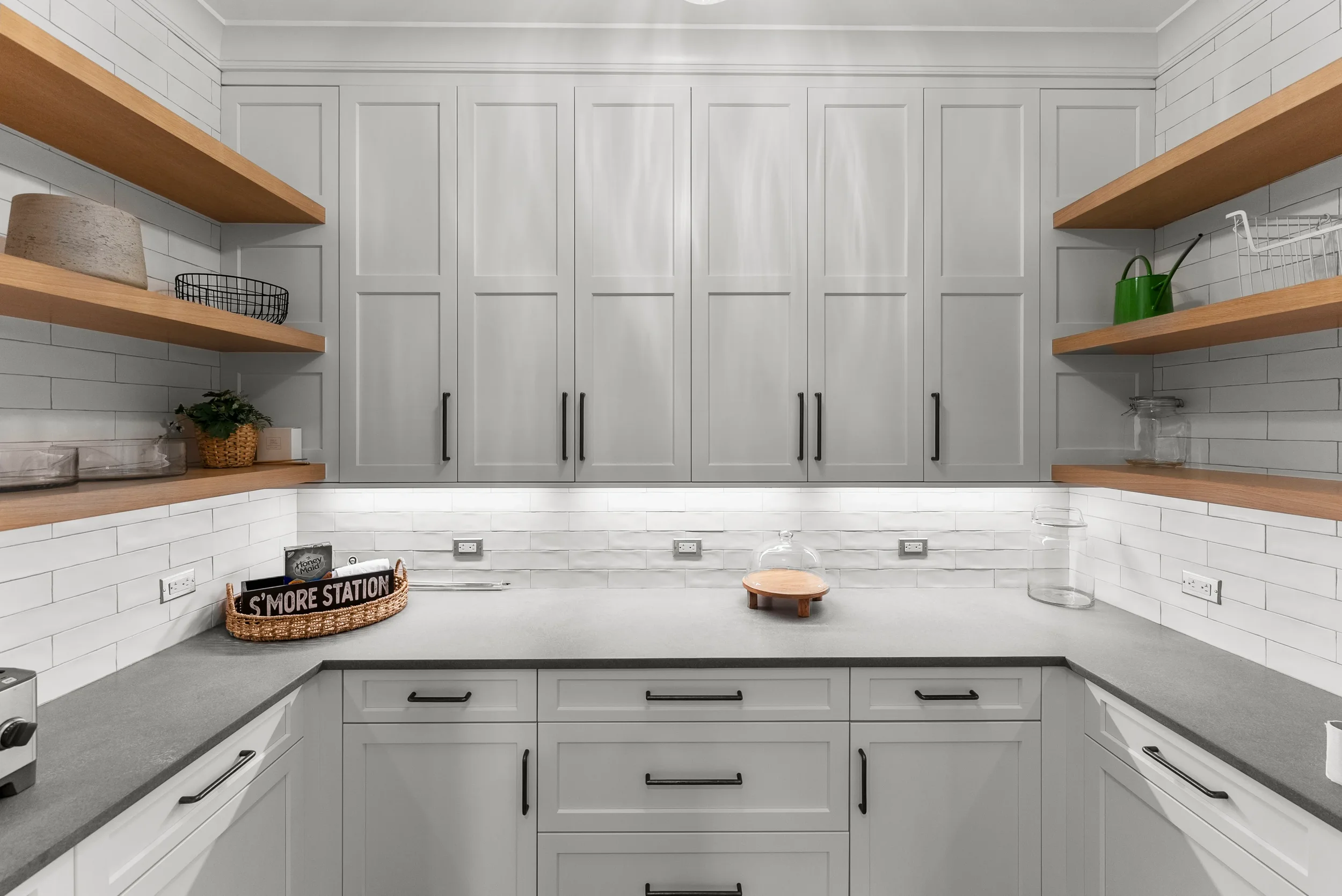 Modern kitchen with white cabinets, gray countertops, open wooden shelves, white subway tile backsplash, and various kitchen items such as a green watering can and glass jars.