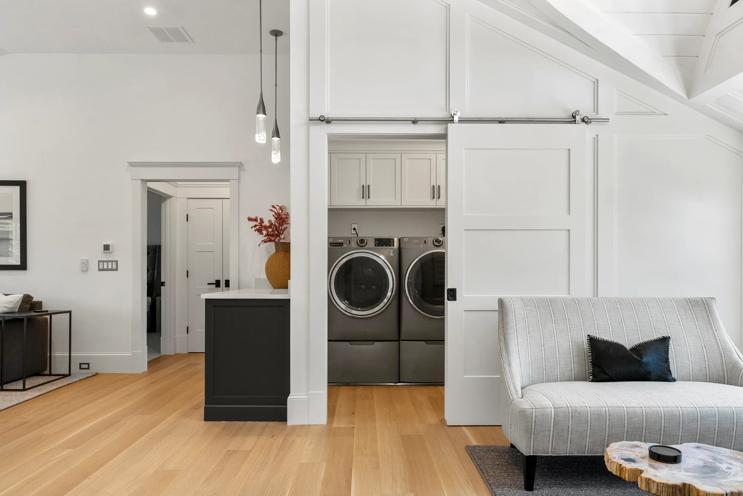 Living room with white walls, wood flooring, a gray and black loveseat, a rustic coffee table, and a laundry area with washer and dryer behind a sliding barn door.