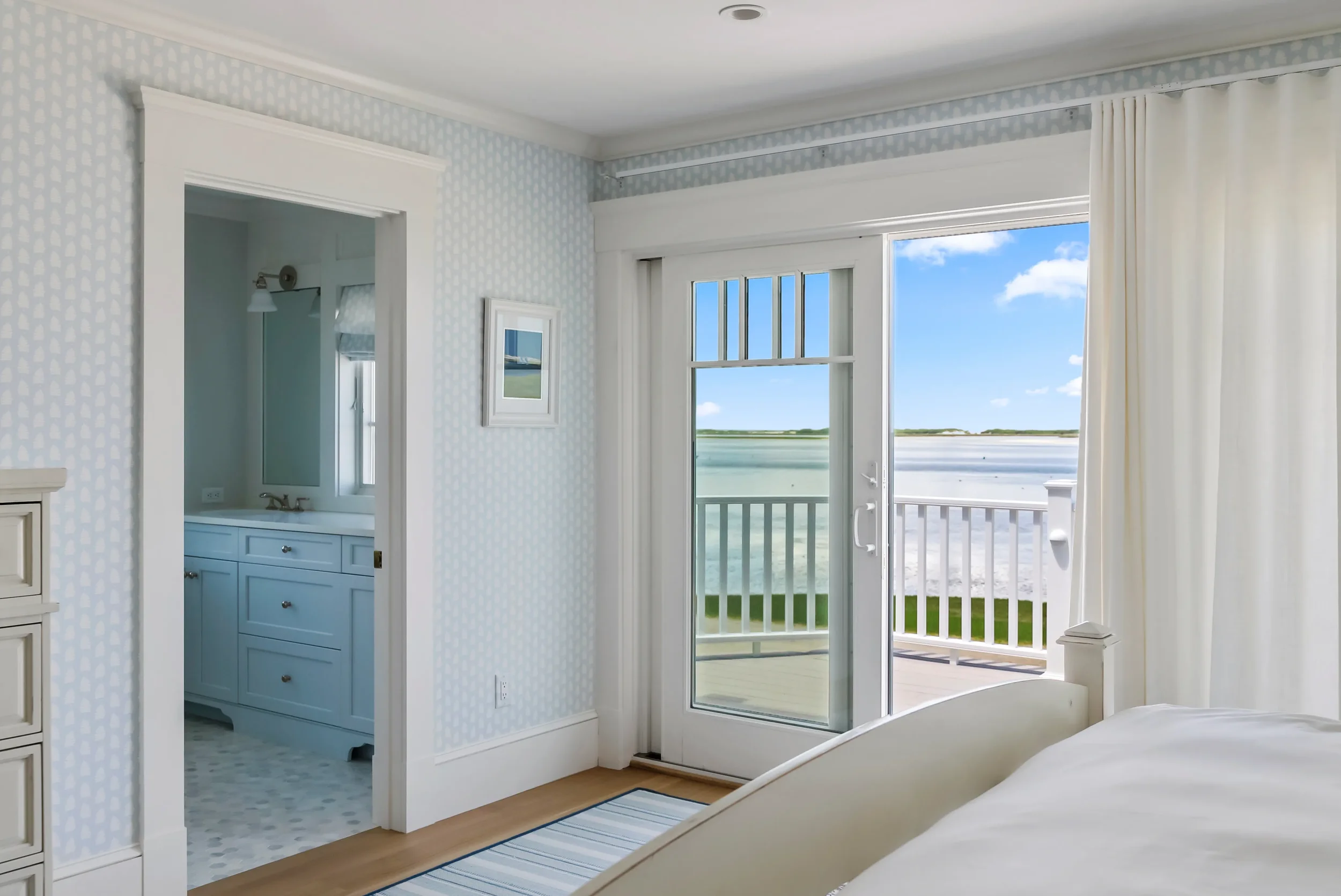 A bedroom with a sliding glass door leading to a balcony, offering a view of water and sky. The room has white furniture, blue patterned wallpaper, and white curtains.