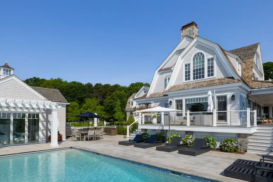 A luxurious house with a swimming pool in the backyard, outdoor seating area with an umbrella, and lush greenery in the background under a clear blue sky.