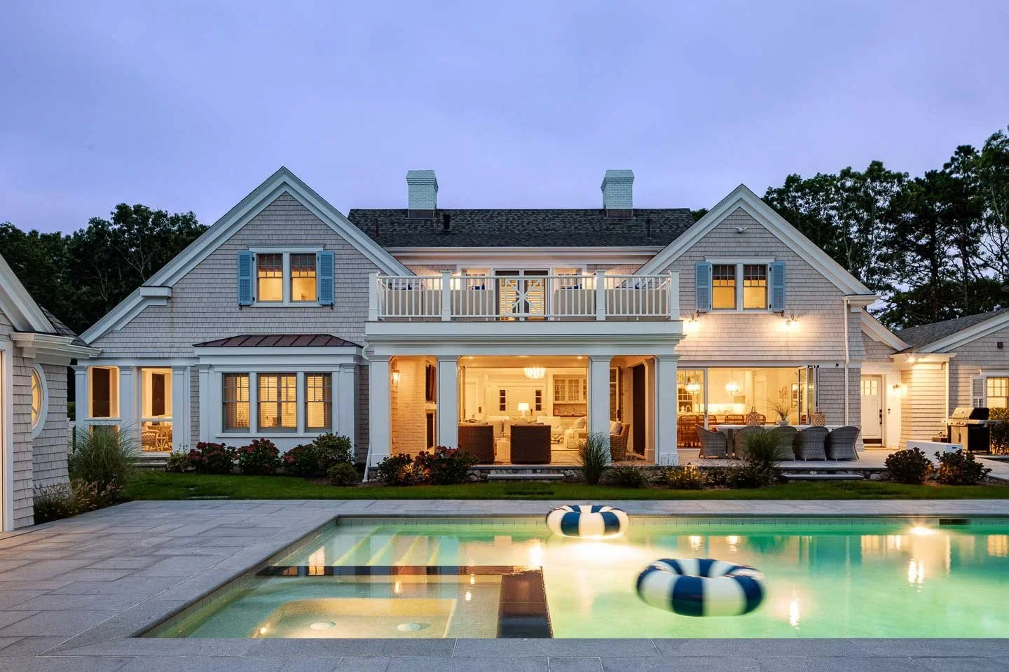 Large two-story house with grey siding and blue shutters, illuminated at dusk, with an outdoor pool in the foreground and landscaped yard.