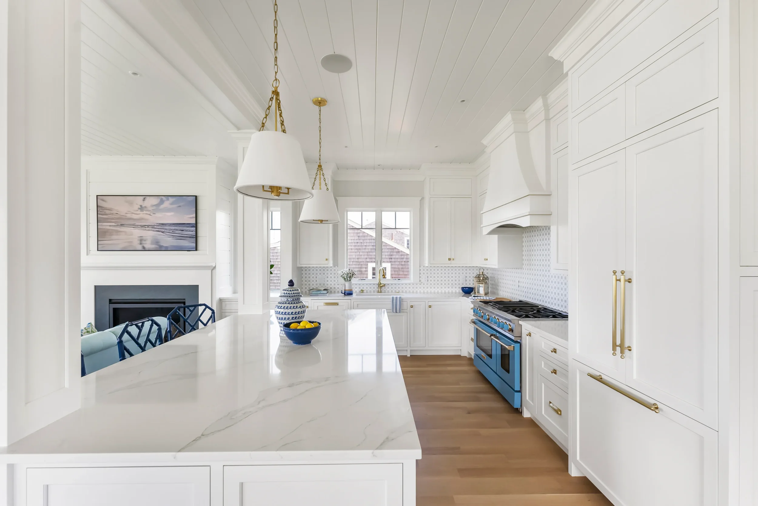 Bright white kitchen with gold accents, blue range, marble island, and window showing a neighboring rooftop.