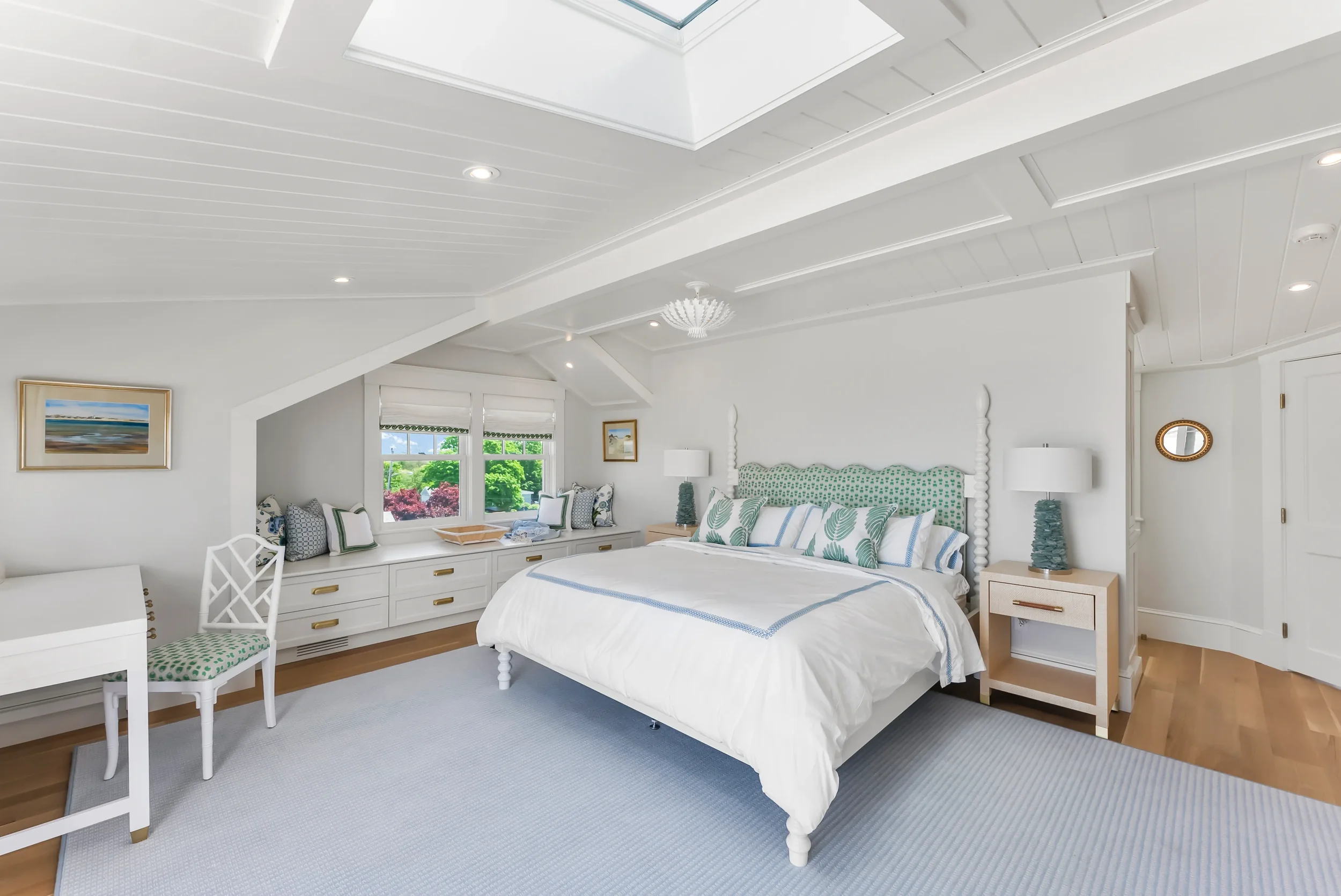 Bright white bedroom with a large bed, nightstands with lamps, a window seat with pillows, and artwork on the walls.