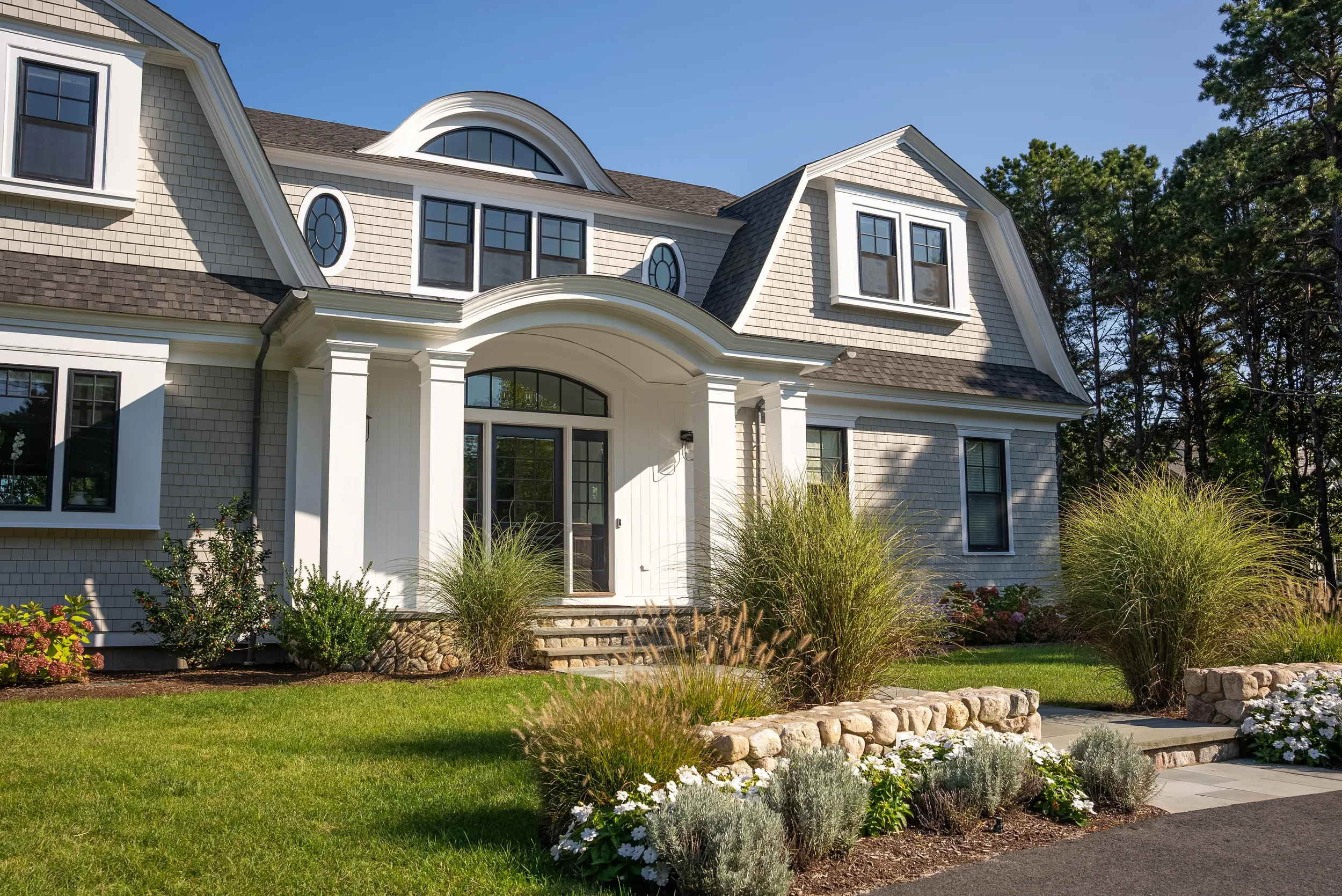 Front view of a large, elegant house with white columns, multiple windows, and landscaped front yard with green grass, plants, and flowers.