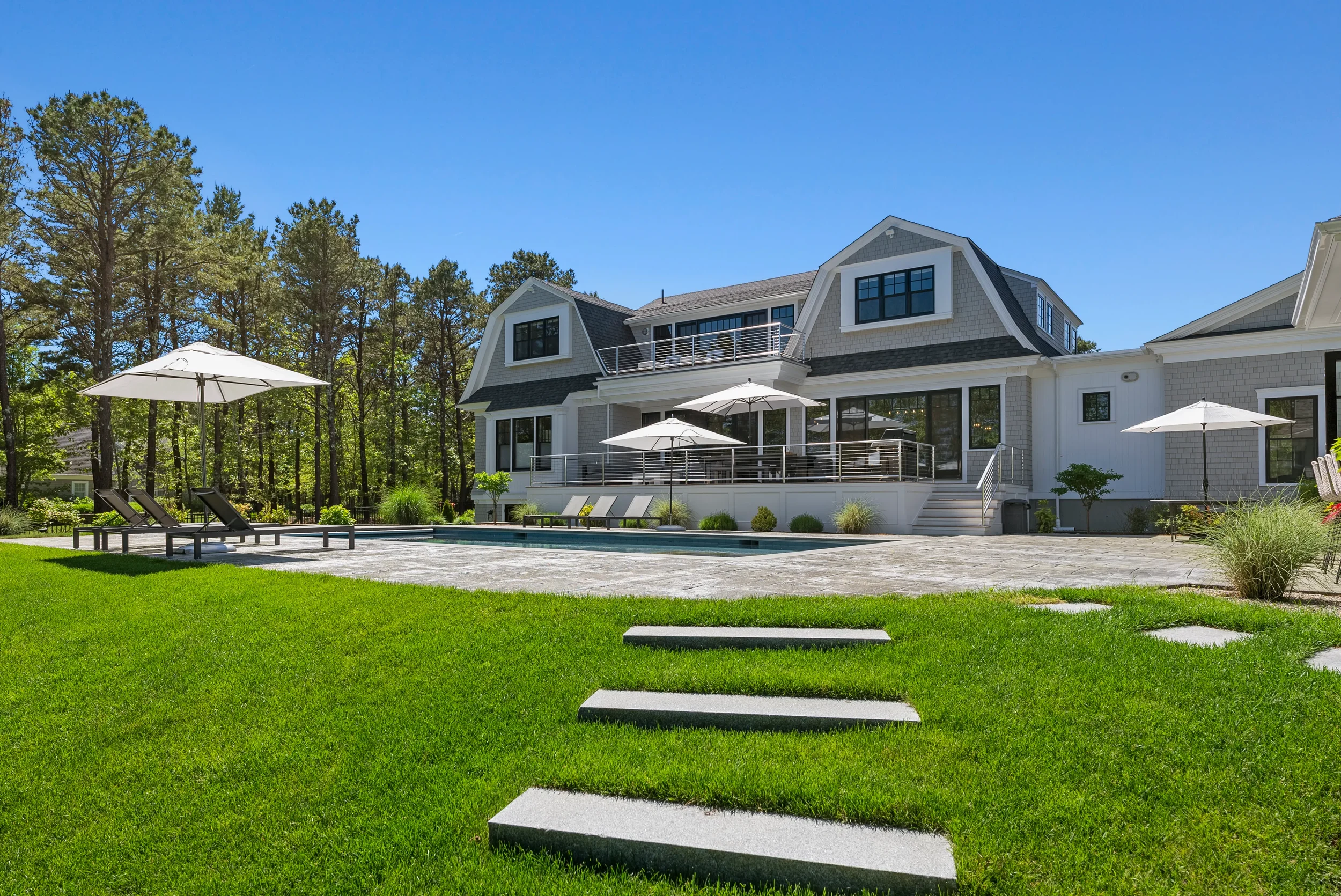 Modern white house with black window frames and front porch, outdoor pool area with umbrellas and lounge chairs, green lawn with stepping stones, surrounded by trees under blue sky.