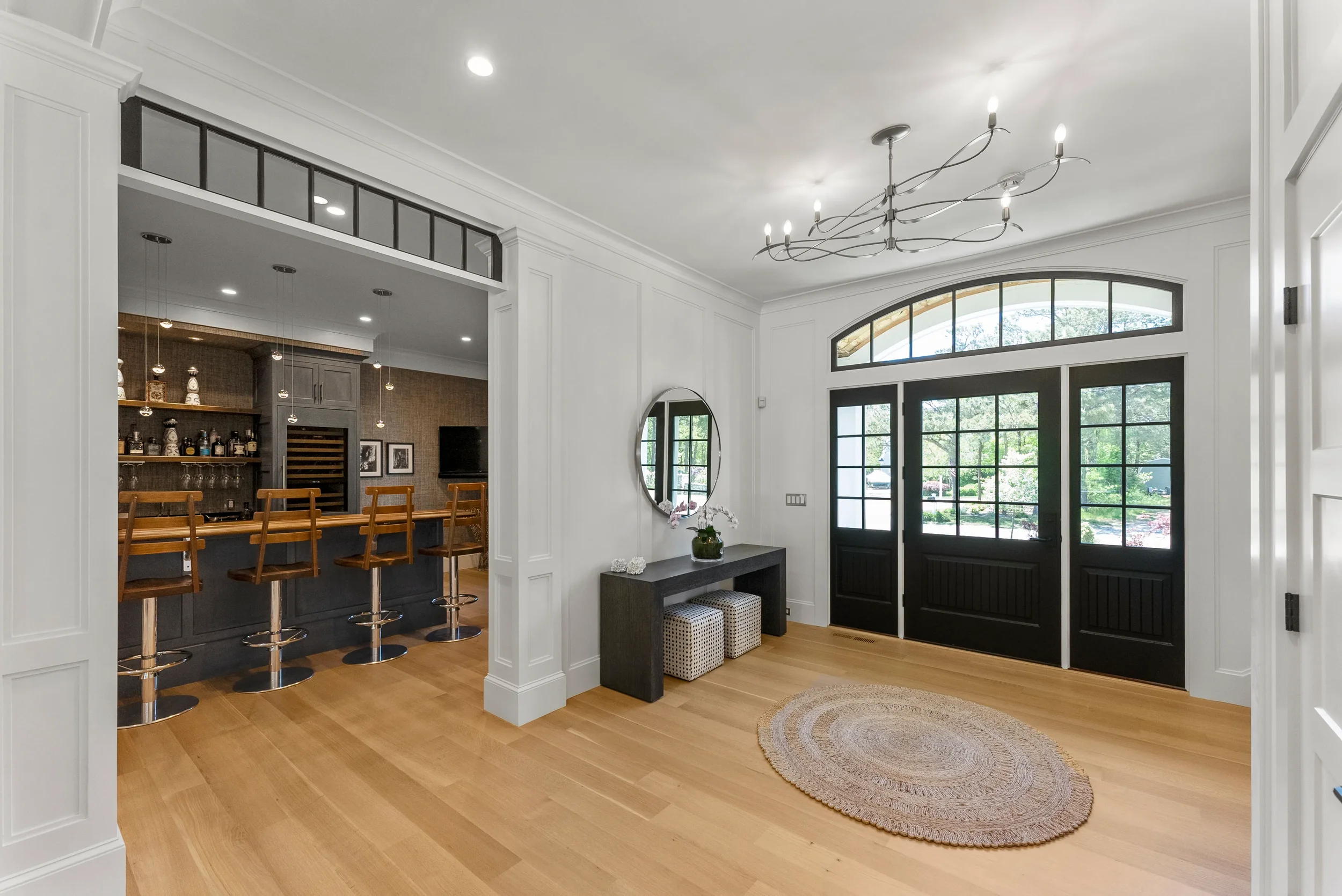 Living room with hardwood floors, black front doors with glass panels, a round mirror, a console table with decorative items, a circular rug, and an entryway leading to a kitchen/bar area.