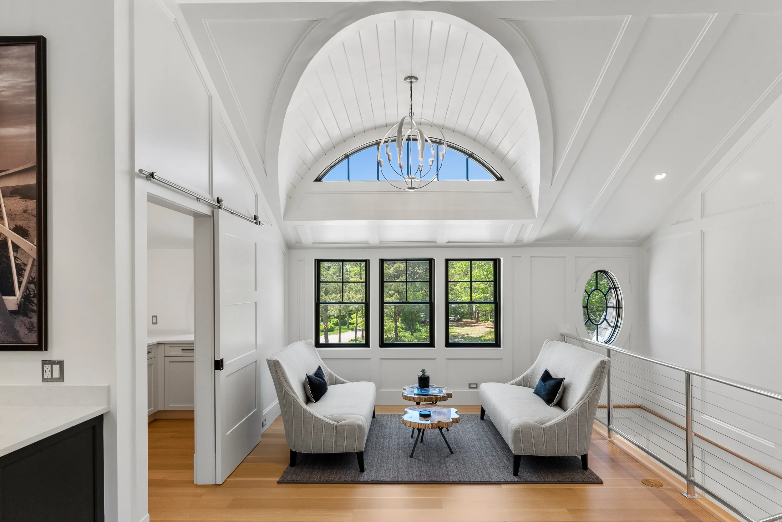 Bright living room with white walls, ceiling, and trim, featuring a central arched ceiling with a modern chandelier, black-framed windows, and two light-colored sofas with black pillows, placed on a gray rug with wooden flooring.