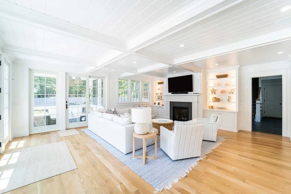 Bright living room with white walls, ceiling, and furniture, featuring a fireplace, television, built-in shelves, and large windows with outside views.