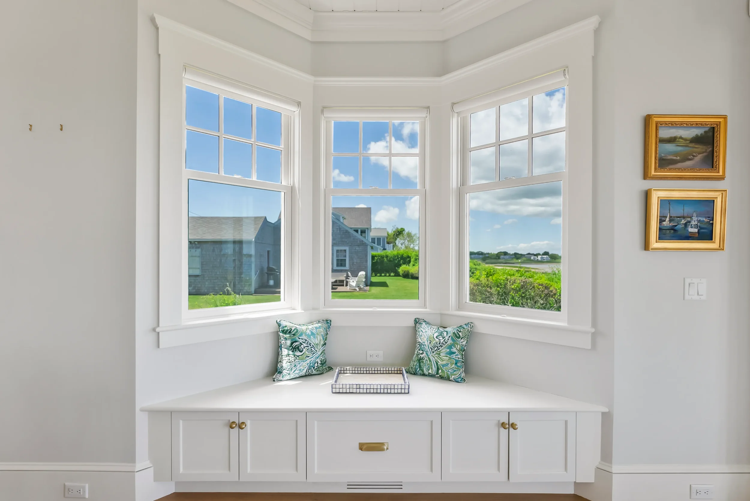 Bright window seat with two decorative pillows and a tray, overlooking a lush green yard and neighboring houses on a sunny day.