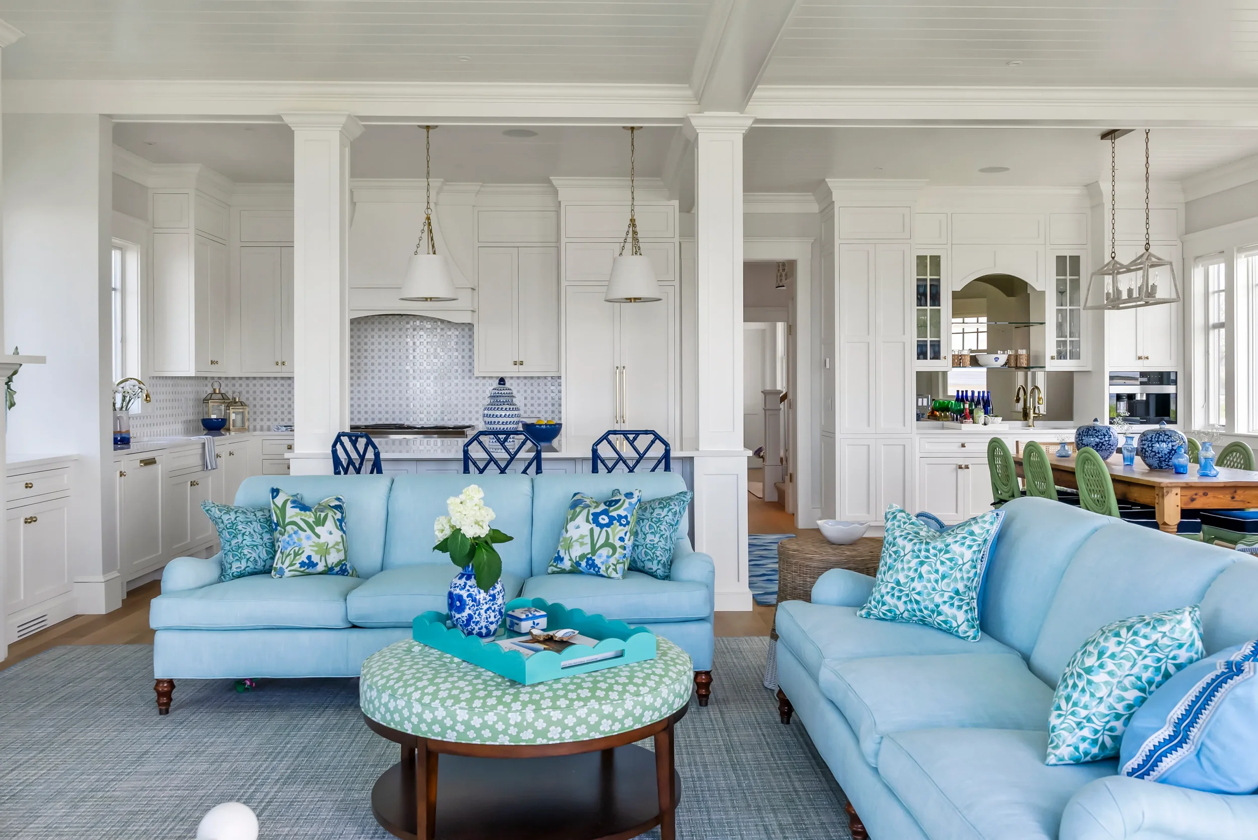 Living room with light blue sofas, floral-patterned pillows, blue vases with flowers, a round green and white ottoman, and a view of a connected kitchen with white cabinetry, blue chairs, and a dining table with blue and green decor.