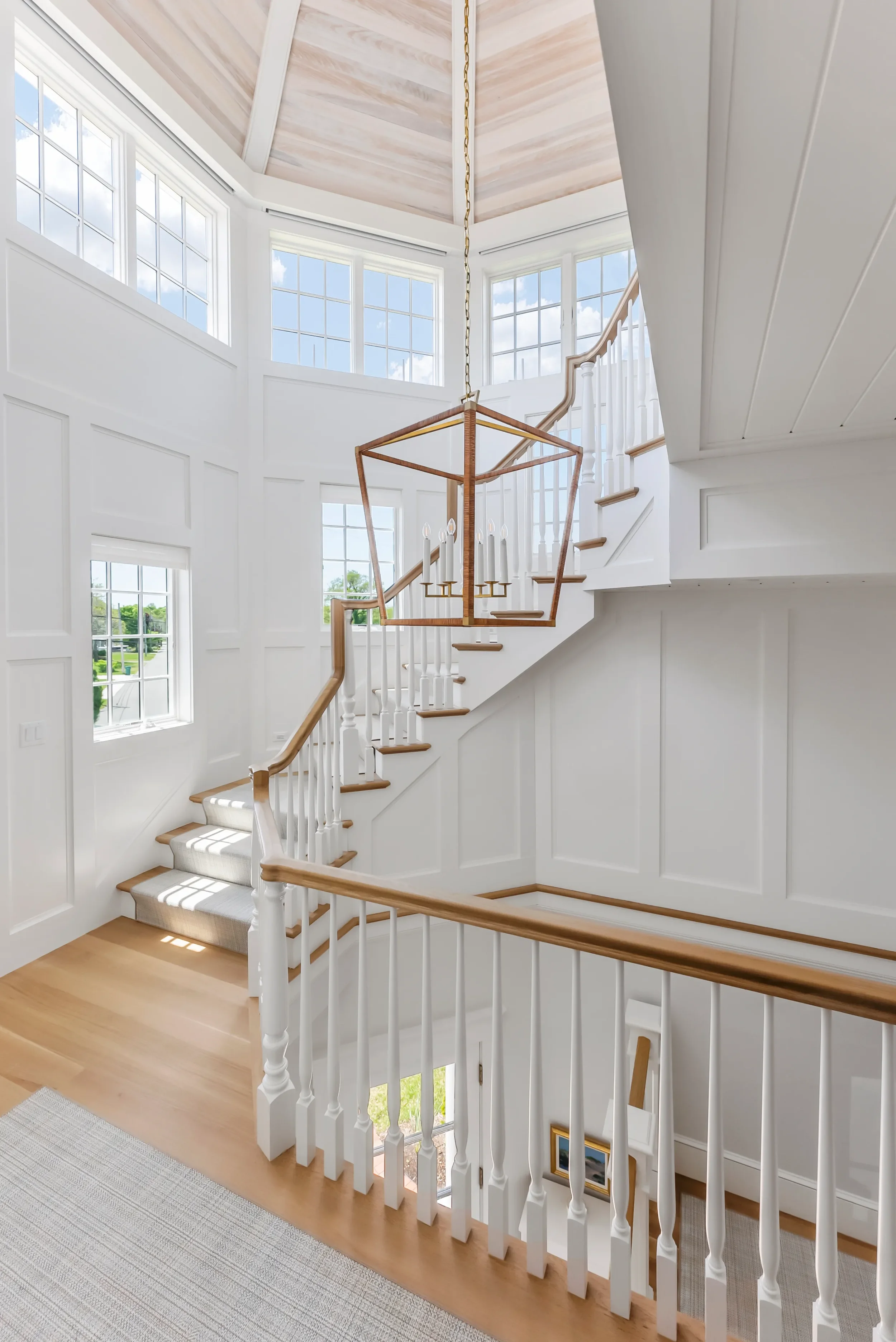 Bright, white multi-story staircase with wooden handrails, a geometric chandelier, and large windows letting in natural light in a modern home interior.