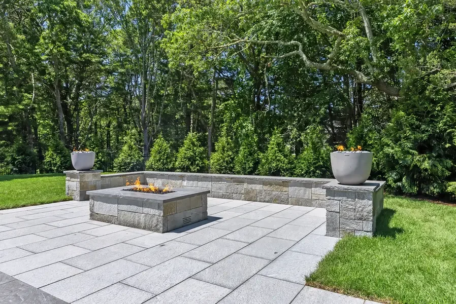 This image showcases a sophisticated outdoor living space featuring a modern fire pit as the central focal point. The rectangular fire pit is constructed with clean-lined stone or concrete materials and displays active flames, creating an inviting ga