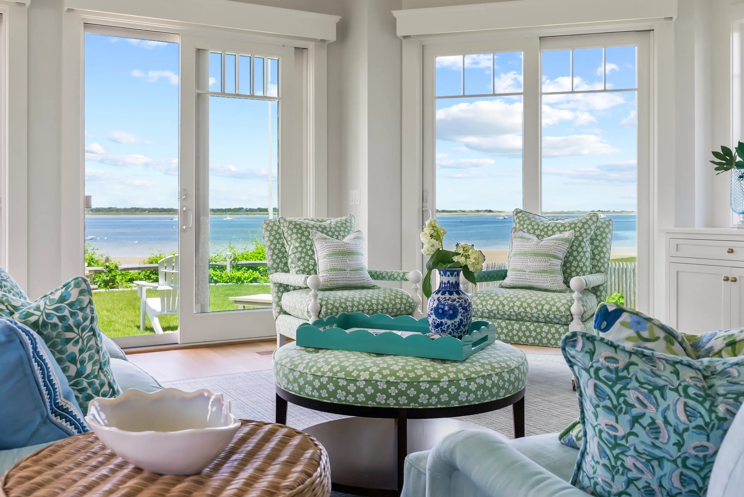 Living room with large windows overlooking a waterfront, green chairs, patterned cushions, and a coffee table with a blue vase of white flowers.