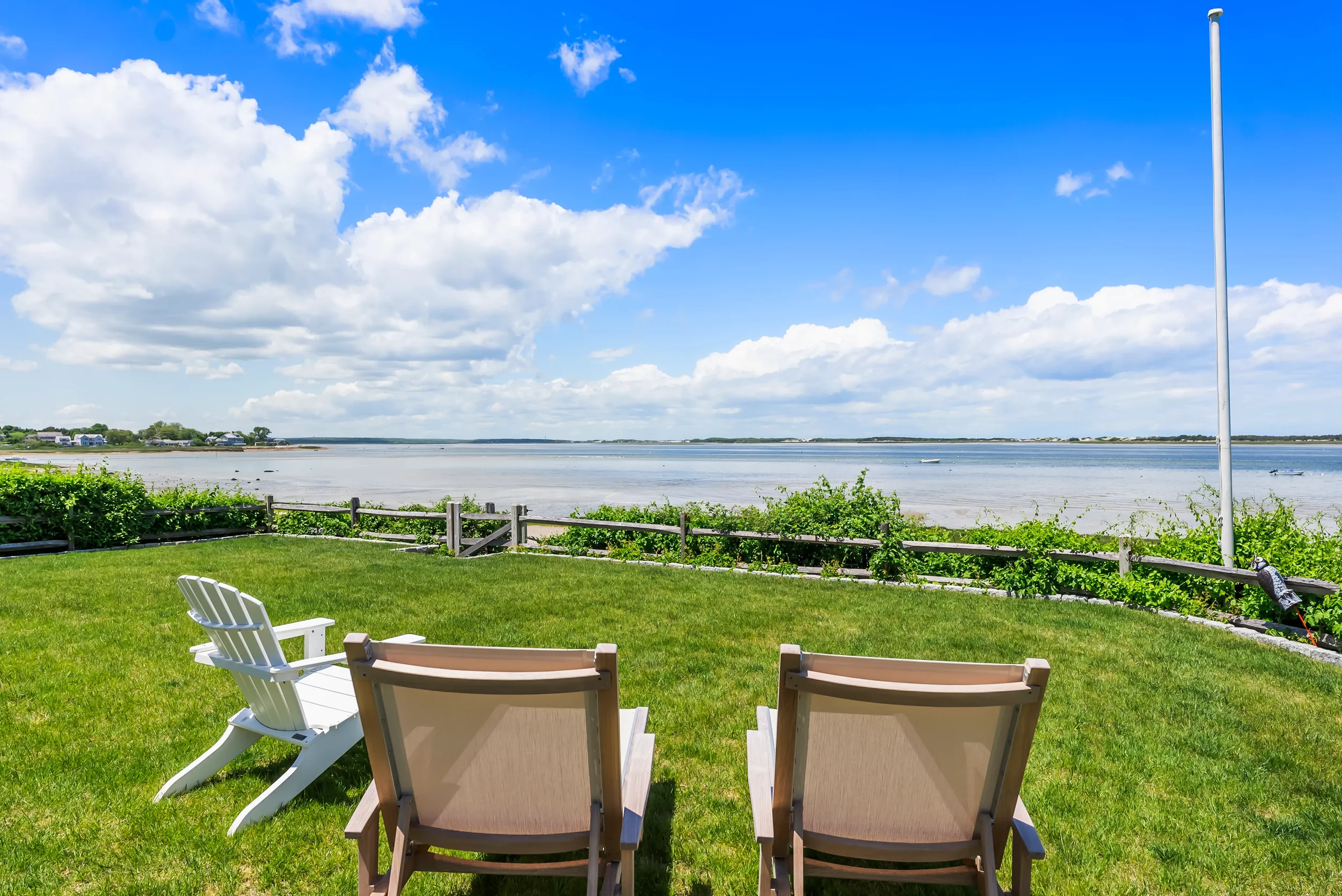 A backyard with three chairs facing a body of water, greenery, and a blue sky with clouds.