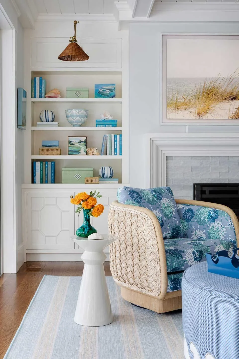 This image captures a cozy coastal-themed living room corner that blends charm, texture, and tranquility. A built-in white bookshelf anchors the space, filled with decorative items like blue-toned books, ceramic vases, bowls, and framed ocean-inspire