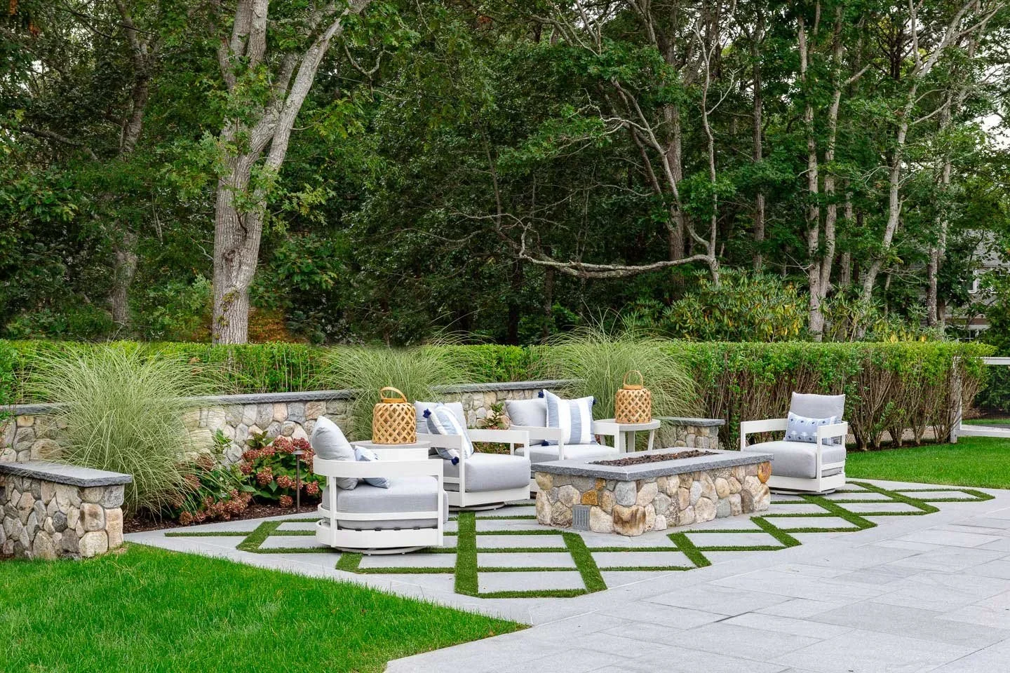 This image showcases a beautifully designed outdoor patio that blends modern comfort with natural elegance. The seating area features sleek white cushioned chairs arranged around a central stone fire pit or table, creating an inviting space for relax