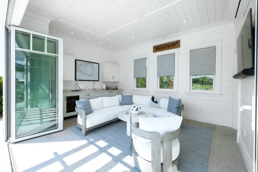 Bright living room with open sliding glass door, white walls, white sofa with blue pillows, small round table, and three windows with shades, overlooking greenery.