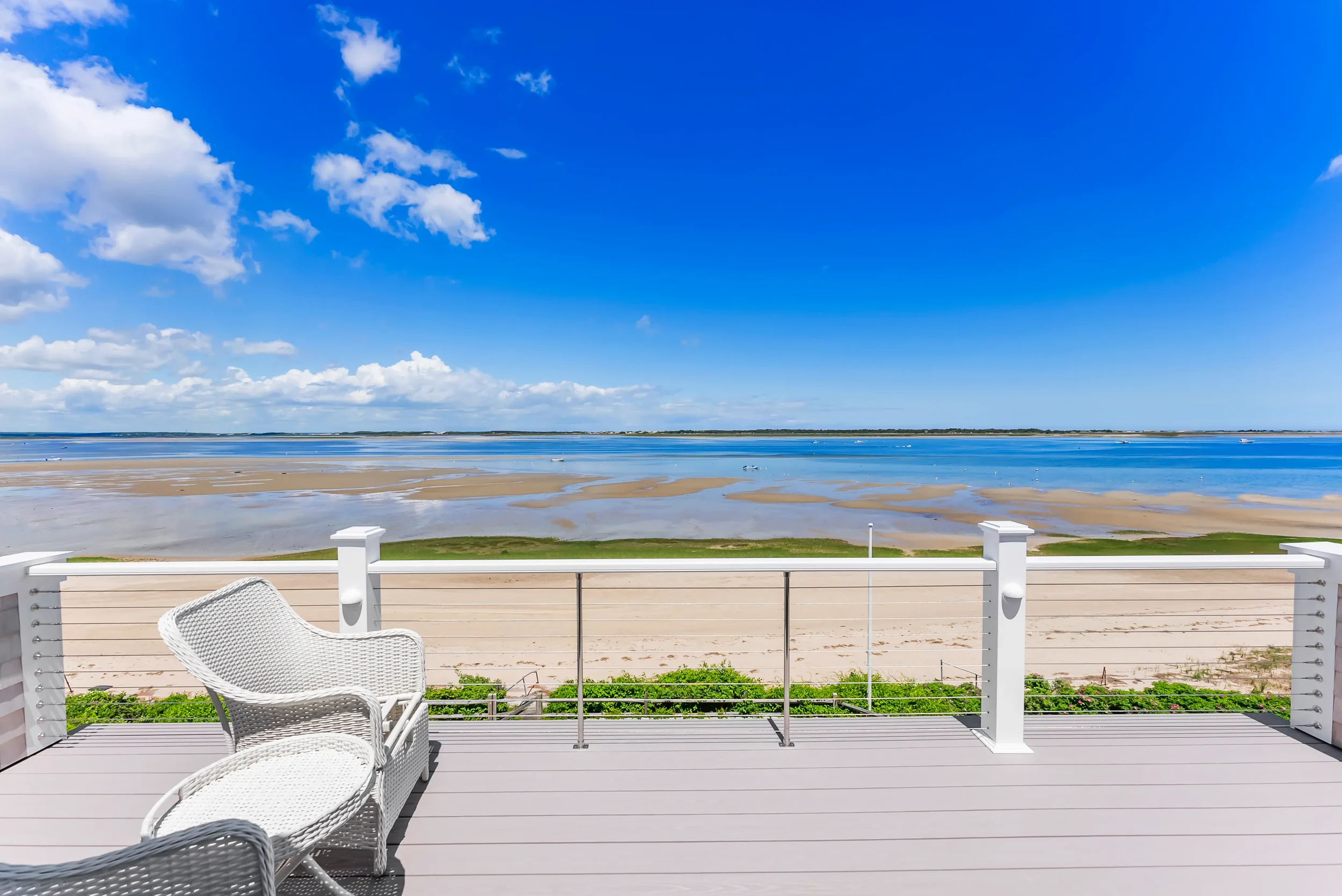 View of a sandy beach and water from a deck with white railing and white wicker chairs under a blue sky with clouds.