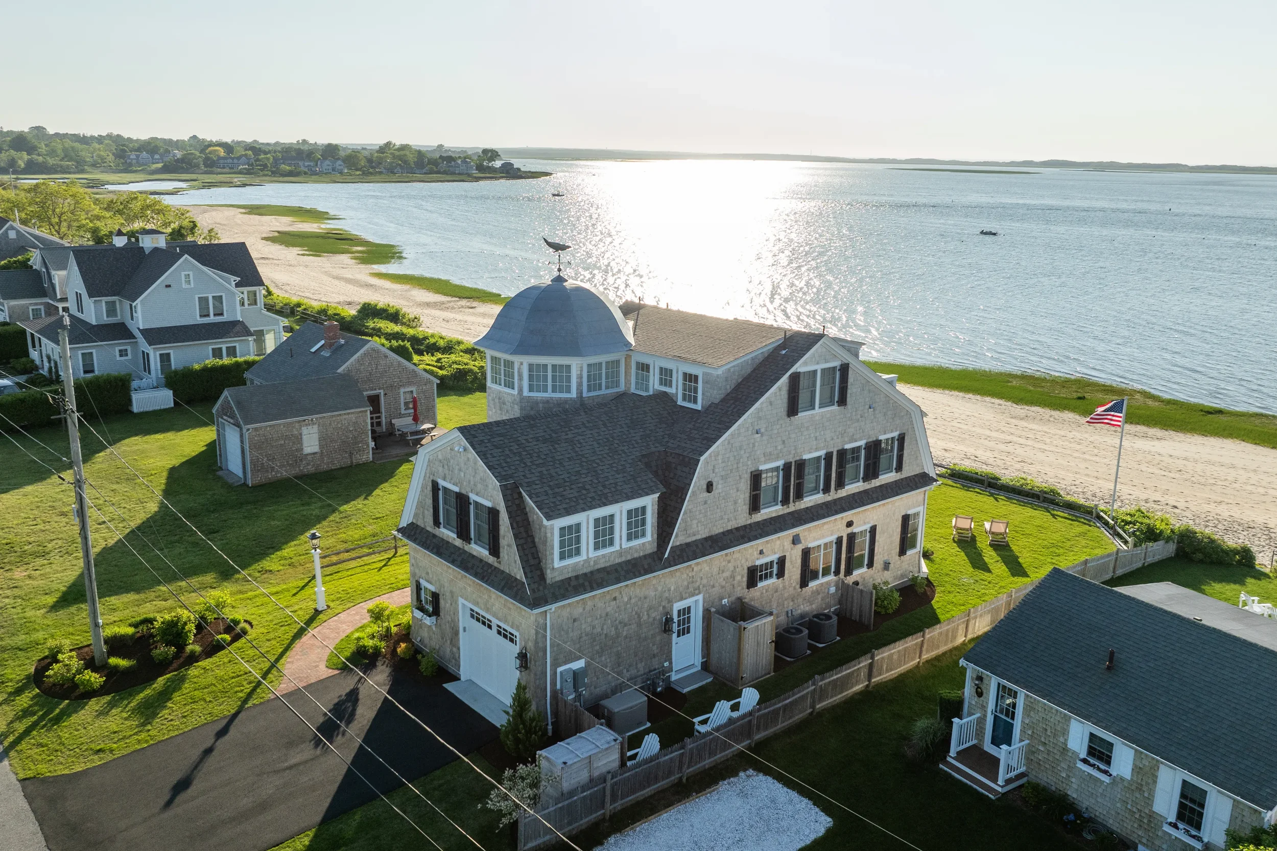 Aerial view of a beach house by the water with a dome roof and flag, neighboring houses, a sandy beach, and boats on the water