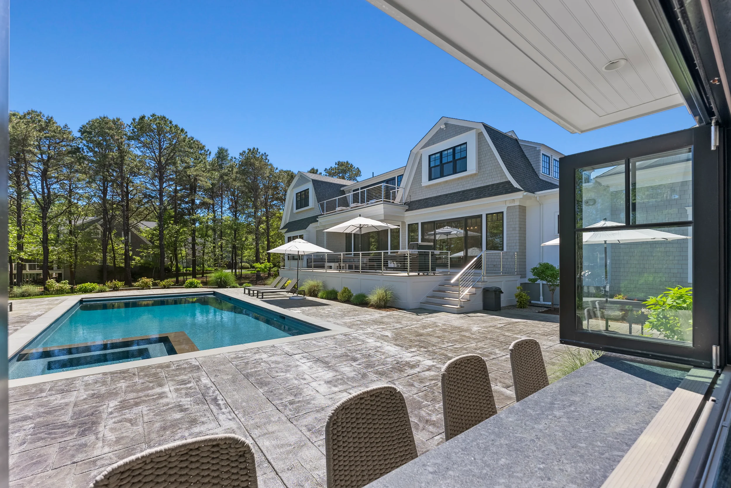 View of a backyard with a swimming pool, patio area with chairs, and a white house with a deck and umbrellas, surrounded by trees on a sunny day.