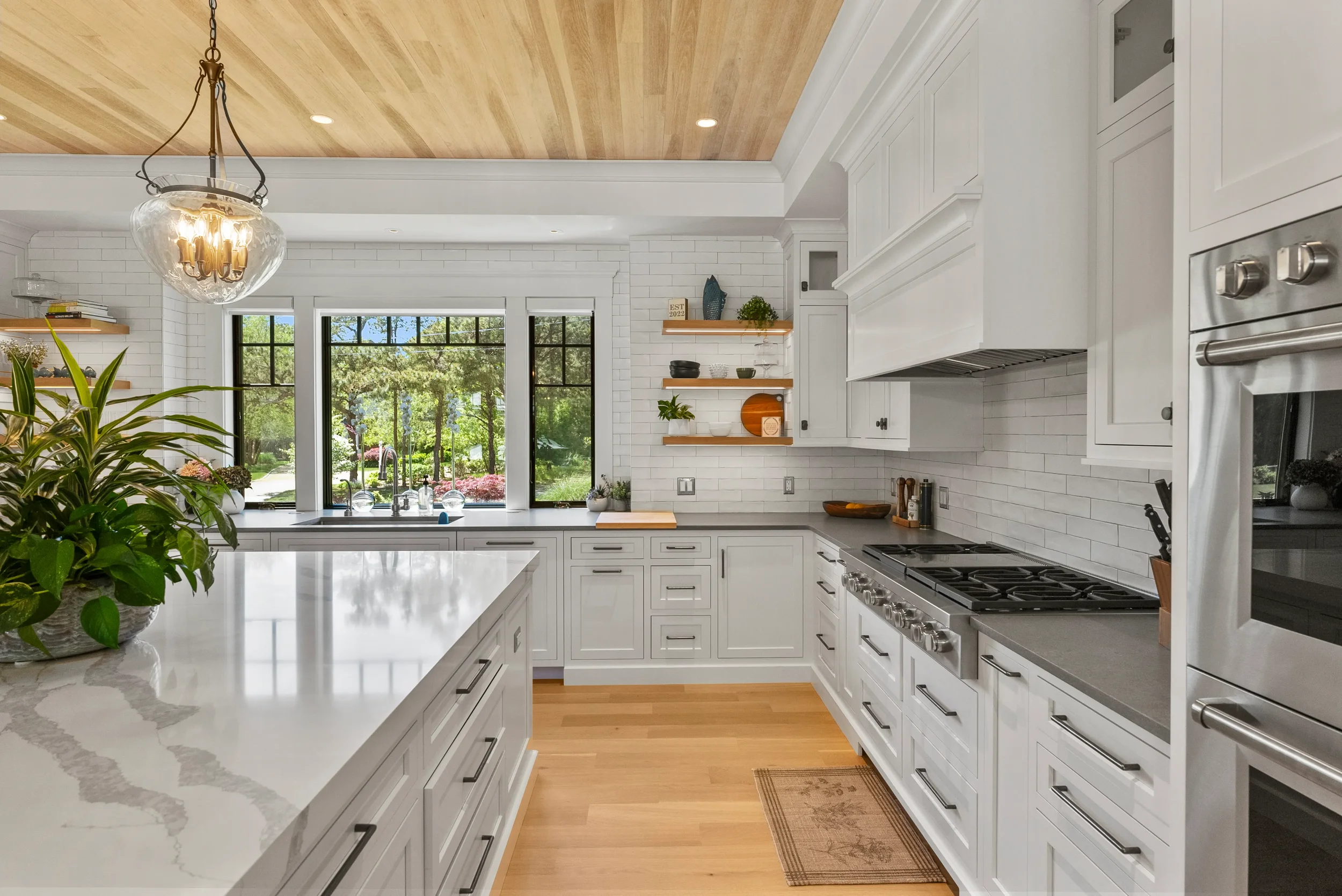 Modern kitchen with white cabinets, gray countertops, wood ceiling, and large window overlooking greenery.