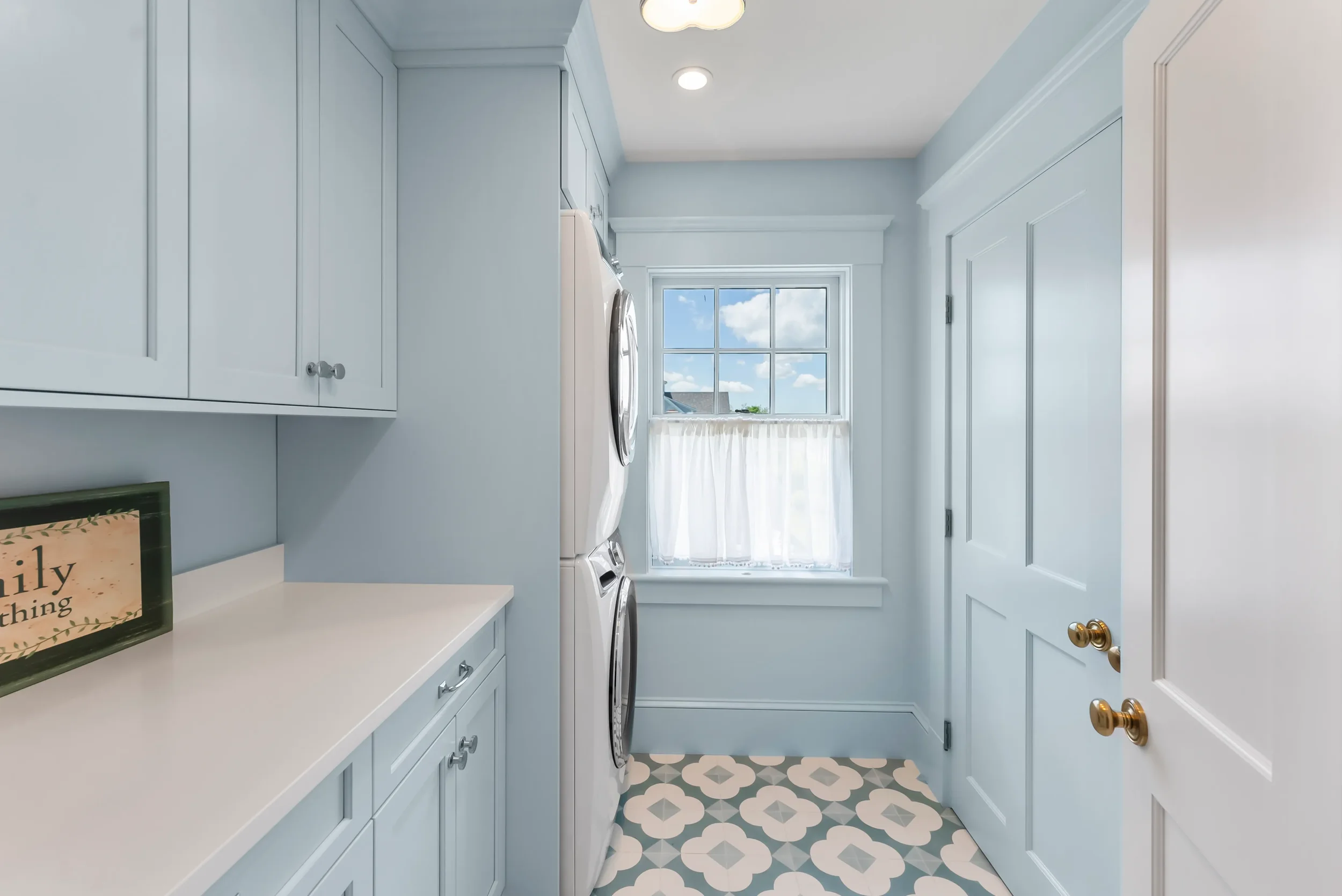 Laundry room with light blue cabinets, white countertop, stacked washer and dryer, window with white curtain and a view of the sky, patterned floor tiles, and a sign that says "family everything."
