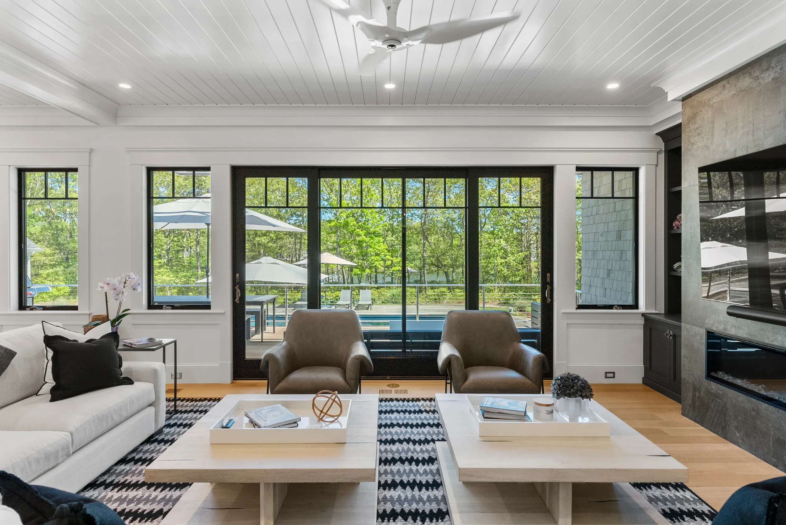 Modern living room with white walls, sliding glass doors leading to an outside porch with patio furniture and umbrellas, two beige armchairs, a white sofa with black pillows, wooden coffee tables with magazines, a patterned black and white rug, and a