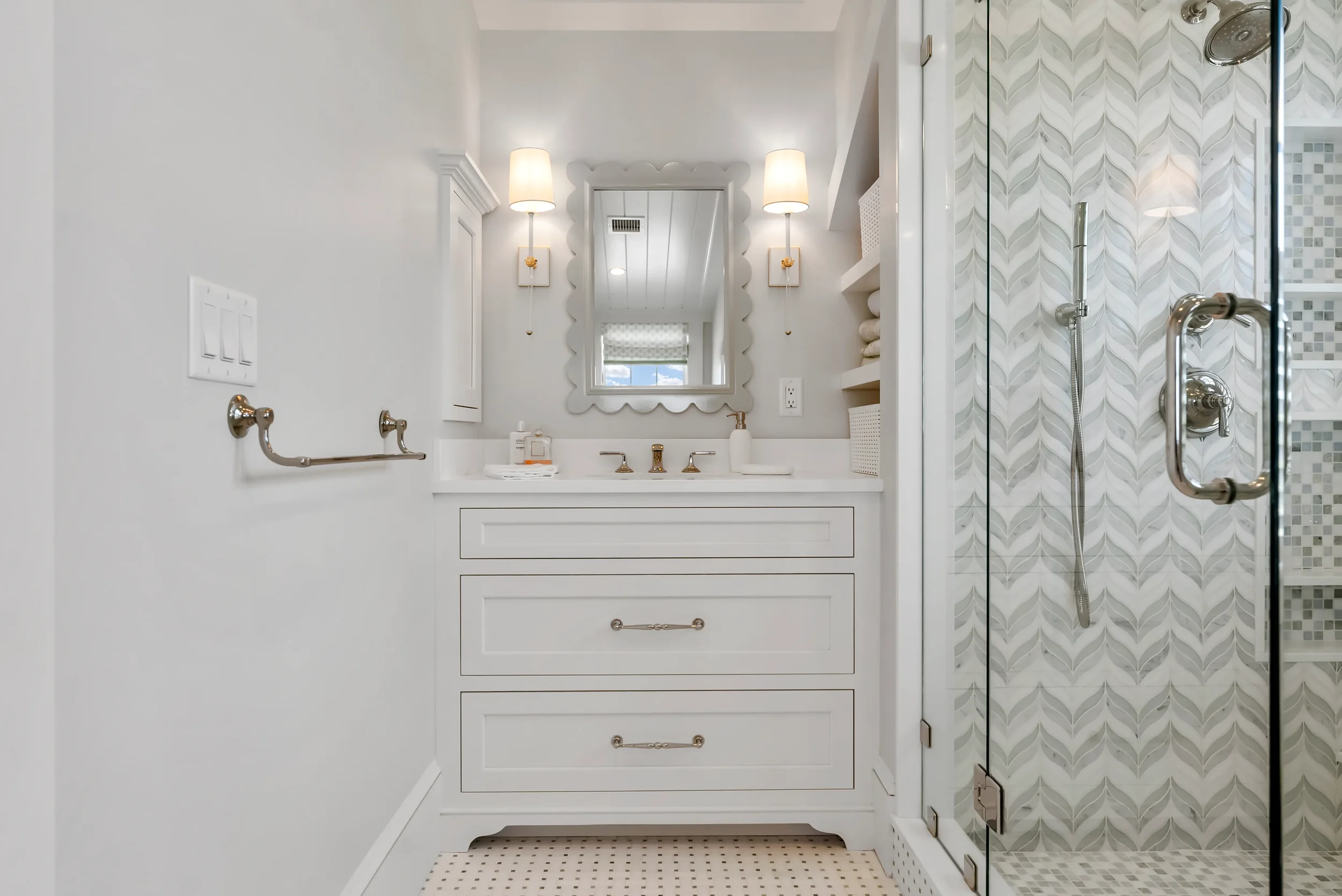 A bright, white bathroom with a vanity, oval mirror, wall sconces, and glass shower enclosure with patterned tiles.