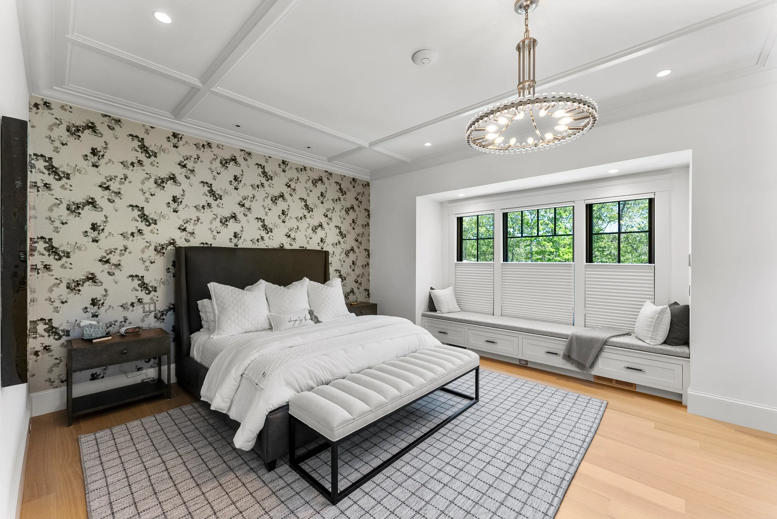 Modern bedroom with white walls and patterned accent wall behind the bed, which has a dark upholstered headboard and white bedding. There are nightstands on either side of the bed, a window seat with pillows below a large window, a circular chandelie