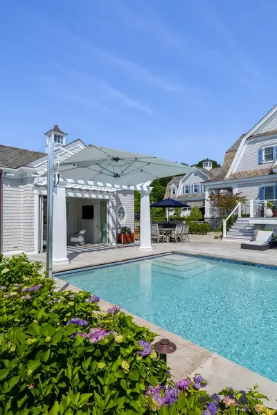 Backyard with a swimming pool, a large white patio umbrella, outdoor furniture, and house buildings with classic architecture under a clear blue sky.