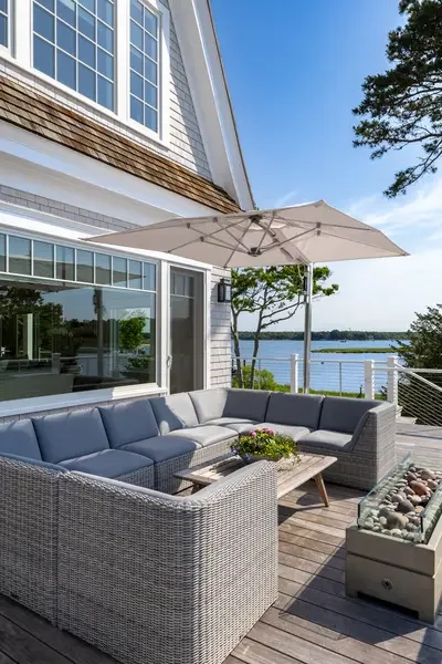 Outdoor patio with gray wicker sectional sofa, table with flowers, large umbrella, next to a house with large windows, overlooking a body of water and trees.