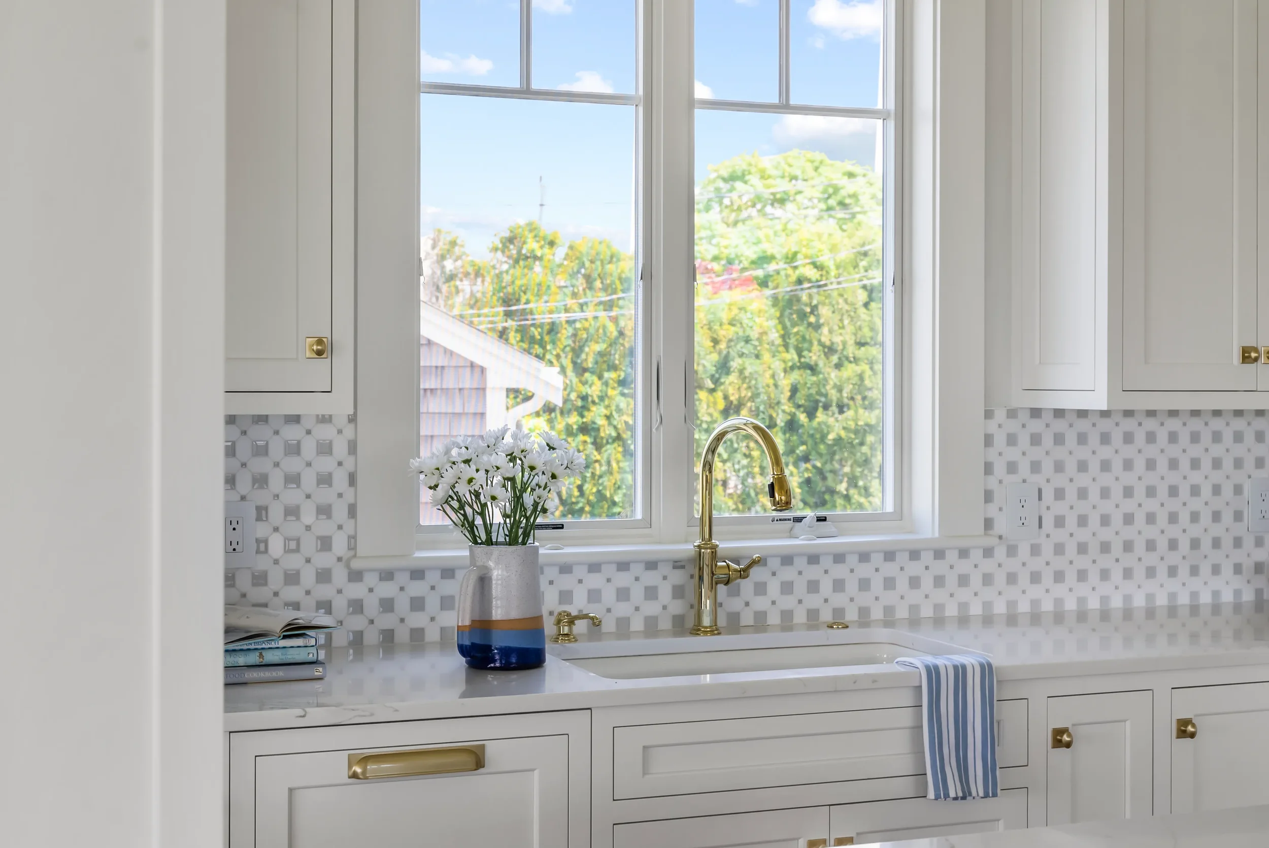 Kitchen sink with gold faucet, white cabinetry, a vase of white flowers, and a window looking outside to trees and a house.