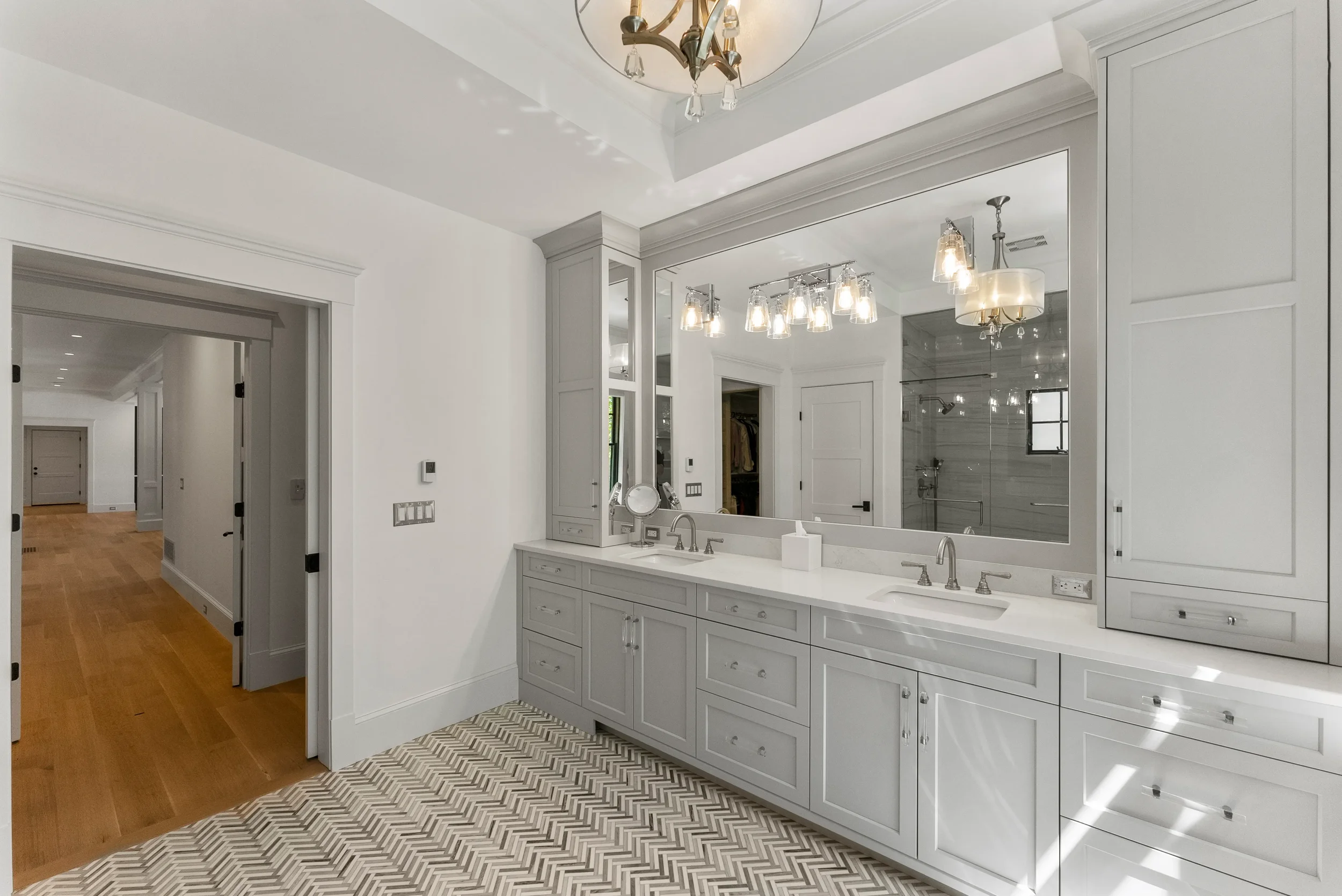 Bright bathroom with white cabinets, large mirror, double sinks, modern lighting, and a walk-in shower with glass door.