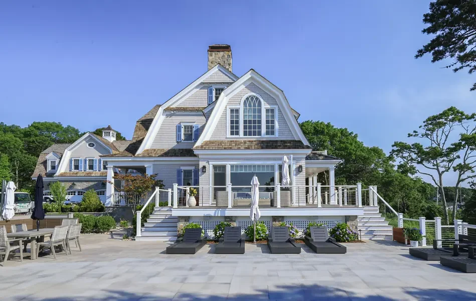 Large white beach house with multiple stories, large windows, and a deck with lounge chairs and umbrellas. Outdoor seating area with dining table and chairs is visible on the patio.