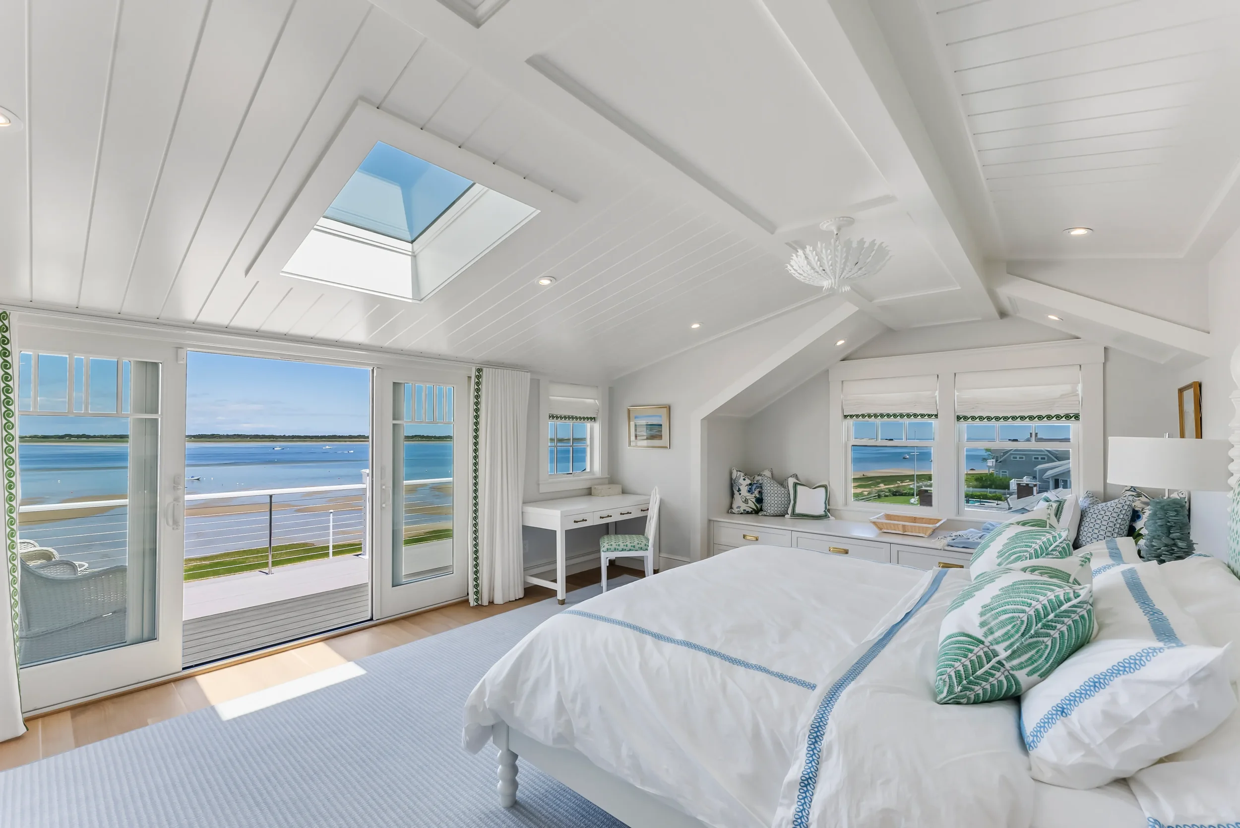 Bright bedroom with a view of water through large windows and a glass door, white walls and ceiling, skylight, decorated with blue and green accents, a white bed with patterned pillows, a small desk, and a chaise lounge under windows.
